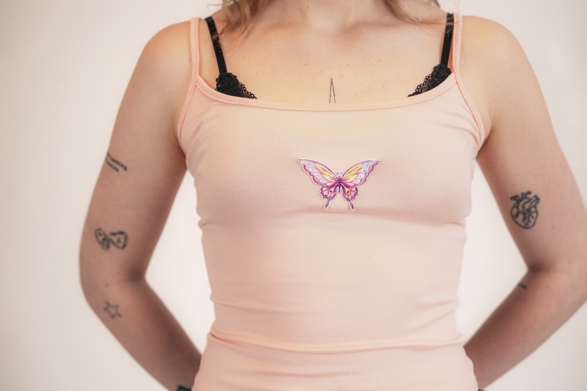 Woman wearing a peach tank top with a butterfly patch on the front, visible tattoos on her arms, and black lace bra straps.