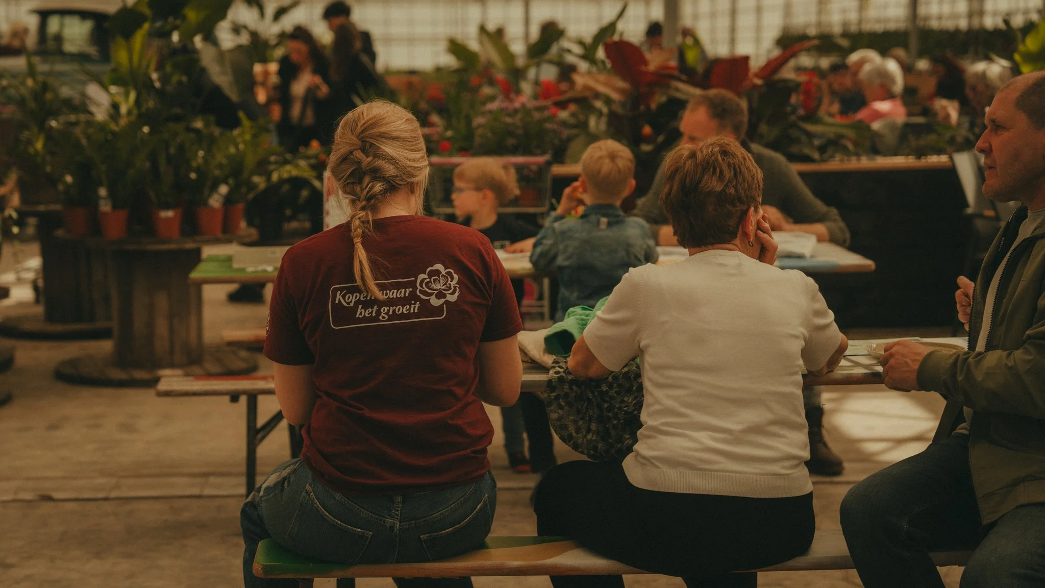 People sitting on benches and tables in a greenhouse or plant shop, engaging in conversation, with potted plants visible in the background.
