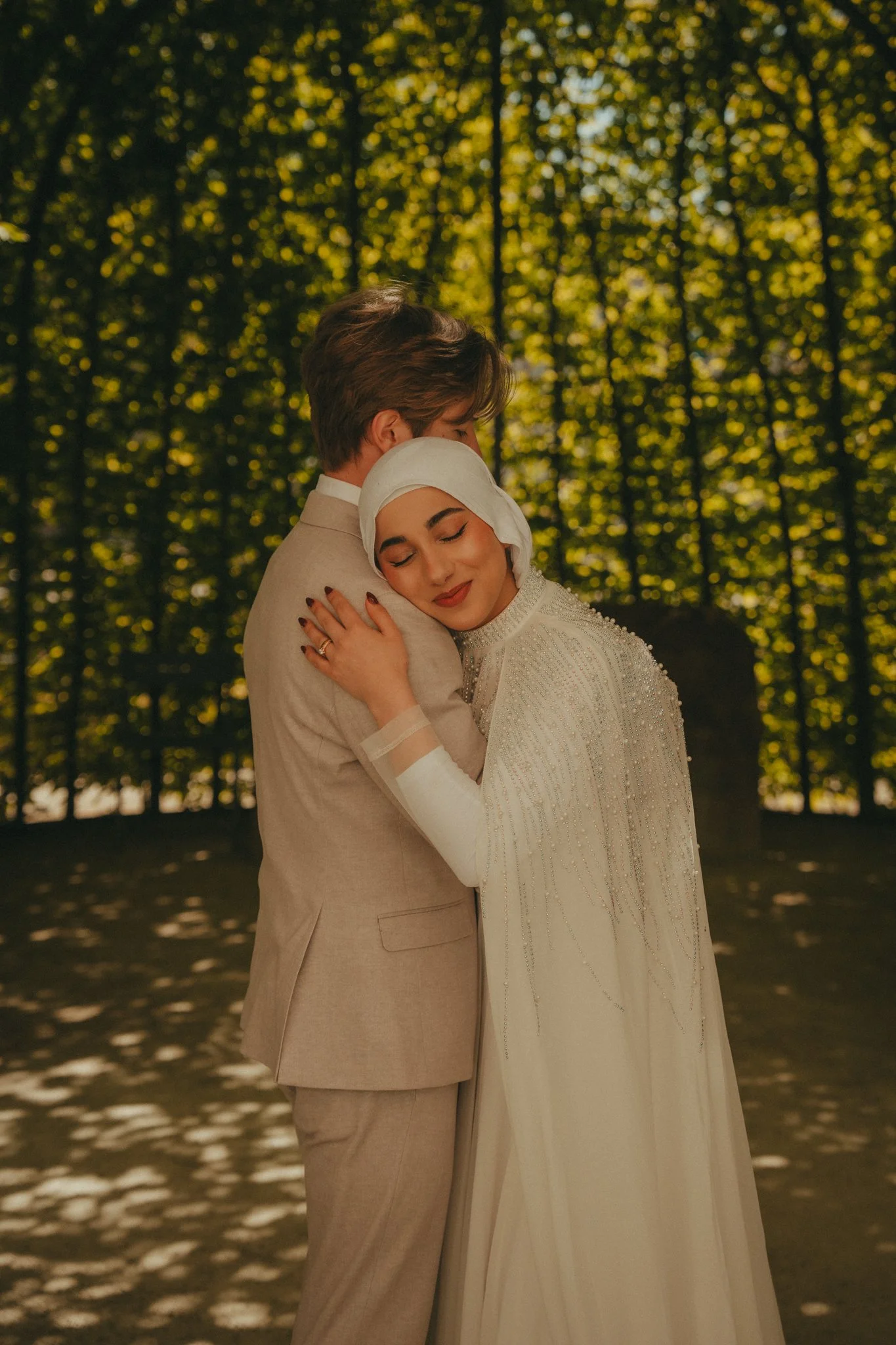 A couple embraces outdoors during sunset with golden leaves in the background. The woman wears a white dress and a headscarf, and the man wears a beige suit. They look content and peaceful.
