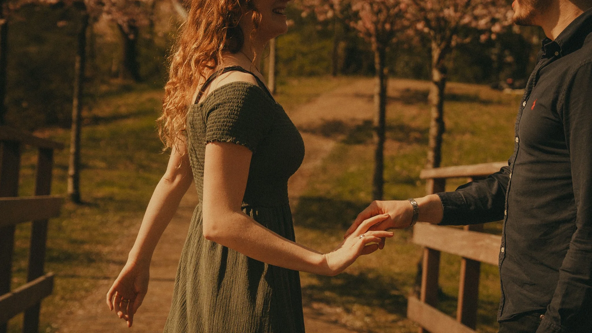 A woman and a man are holding hands and facing each other outdoors during the evening. The woman has curly red hair and is wearing a dark green dress, while the man is wearing a black shirt. There are trees and a fence in the background.