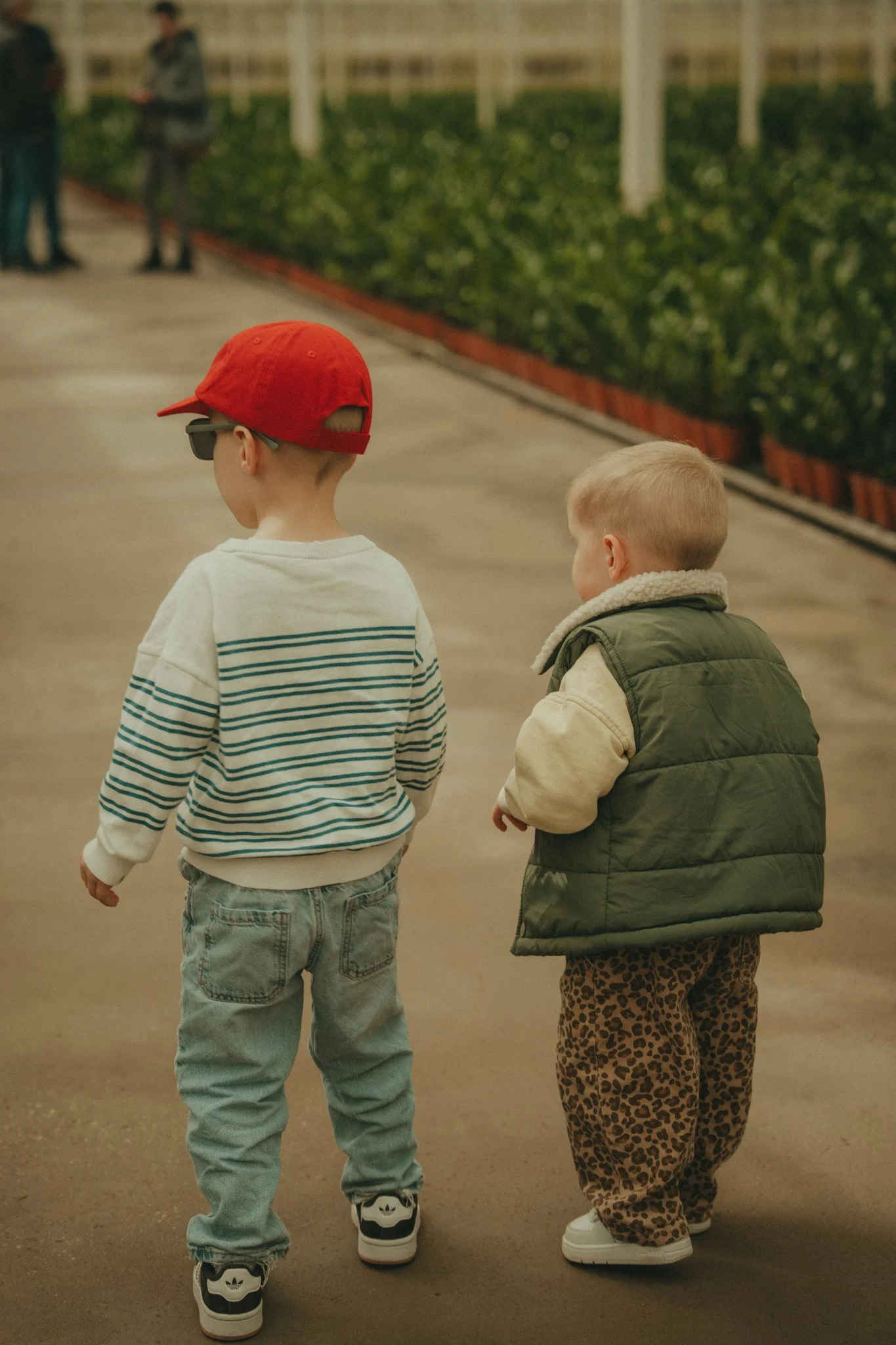 Two young children walking outdoors, one wearing a white striped sweater, a red cap, and sneakers, while the other wears a green quilted vest and leopard-print pants.