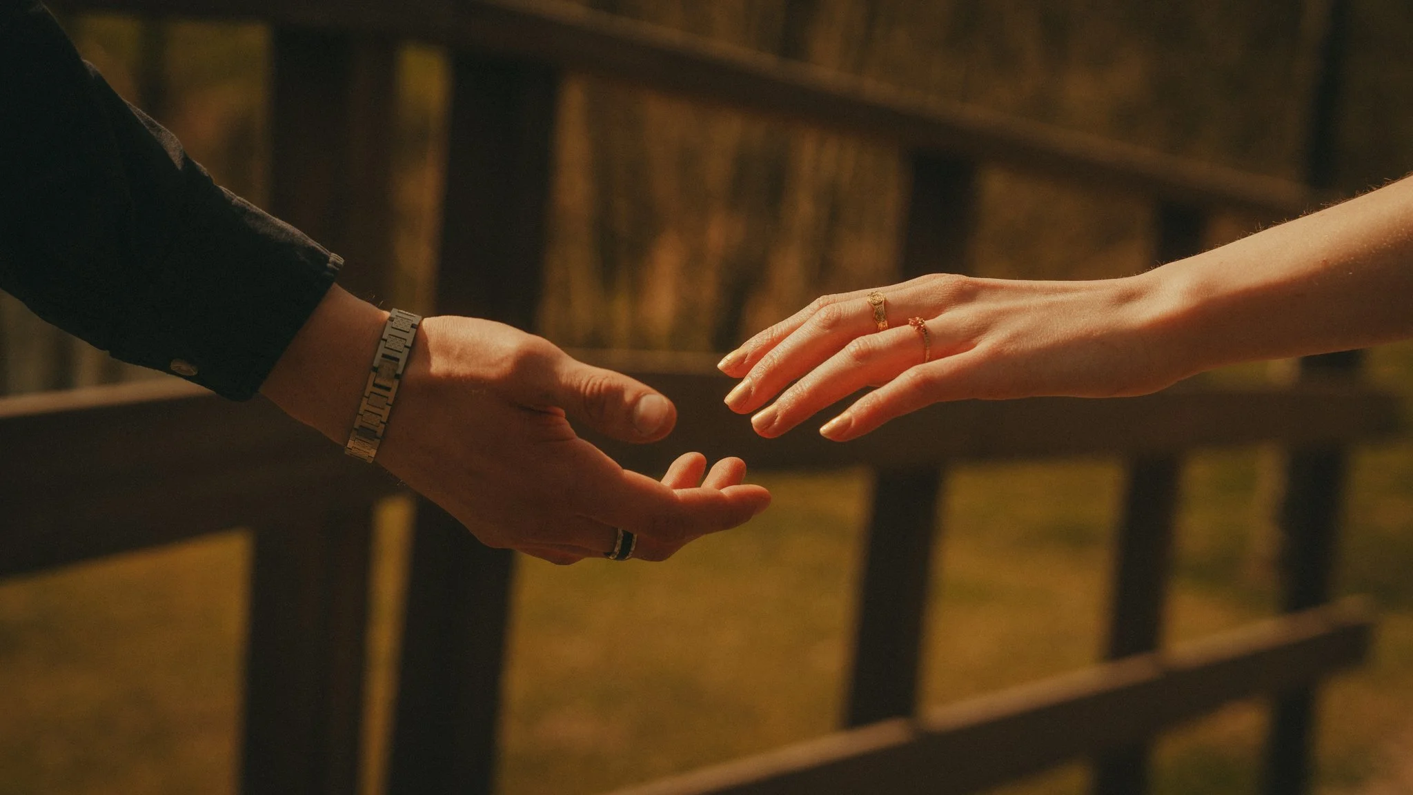 Close-up of two hands reaching towards each other, one with a watch and bracelet, the other with rings, set against a wooden fence background at night.