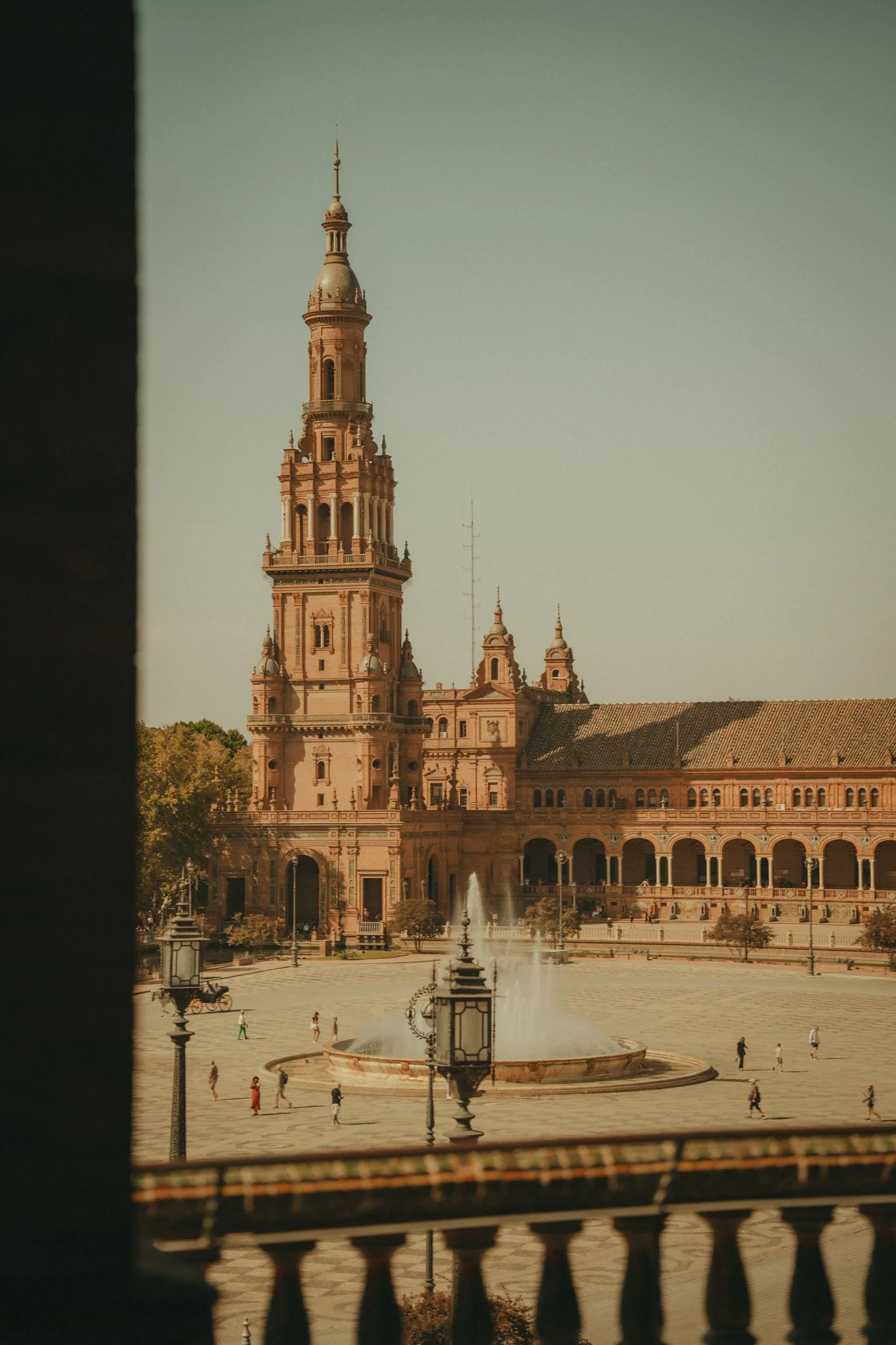 View of a historic building with a tall clock tower, fountains, and a large open plaza with some people walking around, seen through a balcony railing.