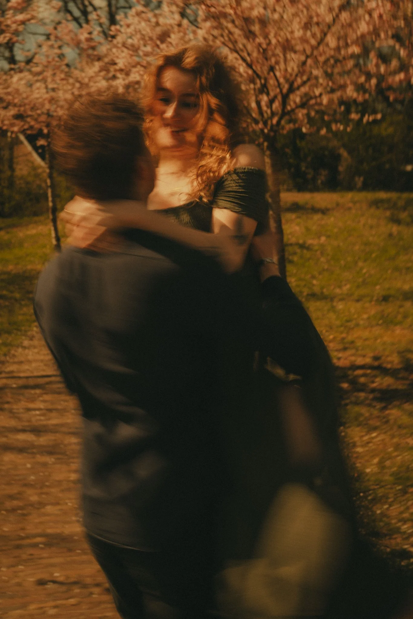 A woman with curly hair embracing another person outdoors at night, with pink flowering trees in the background.