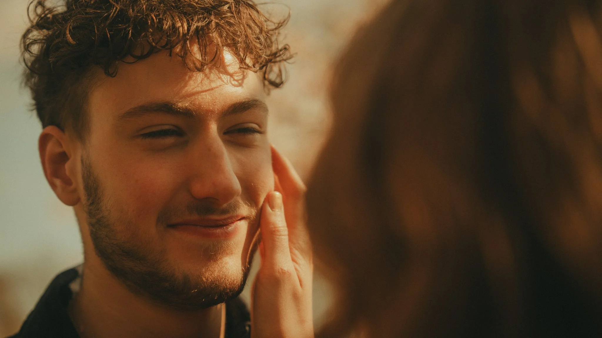 A young man with curly brown hair, light skin, and facial hair is smiling while a person gently touches his face.