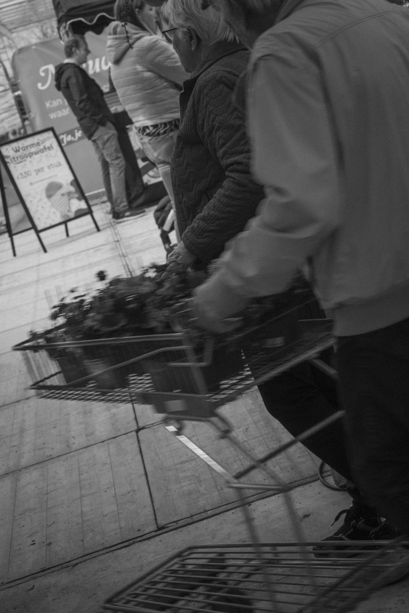 People shopping at a supermarket, one person pushing a shopping cart filled with groceries in the foreground, other shoppers in the background, signs and displays visible.
