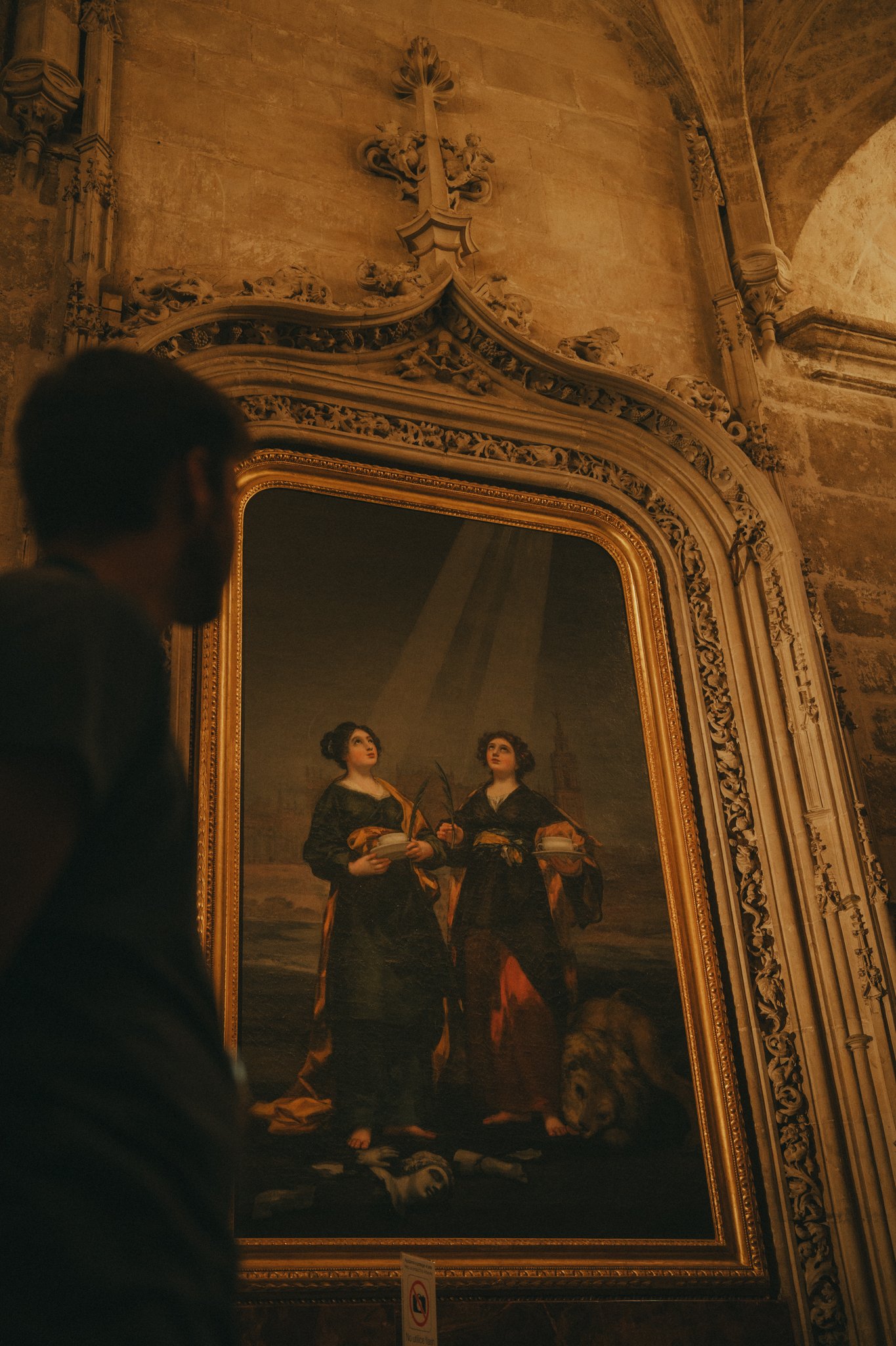 A large framed painting depicts two women standing outdoors, dressed in historical clothing, with a landscape background. The setting appears to be inside a grand, ornately decorated building with stone walls and intricate architectural details.