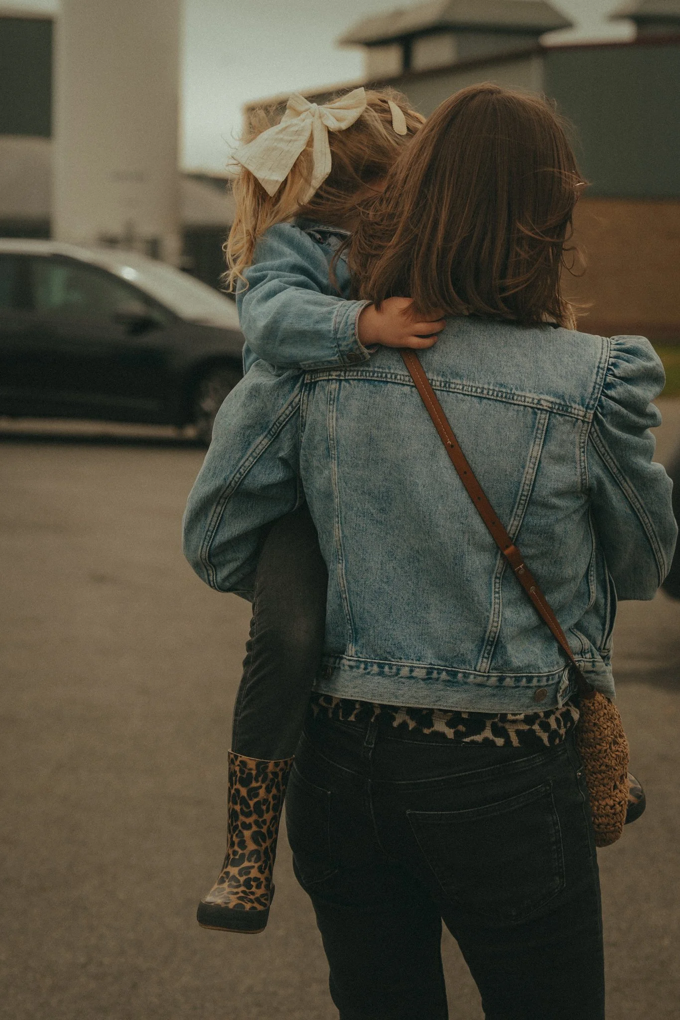 A woman in a denim jacket holding a young girl with curly blonde hair and a large cream bow, wearing leopard print boots, is seen from behind in a parking lot.