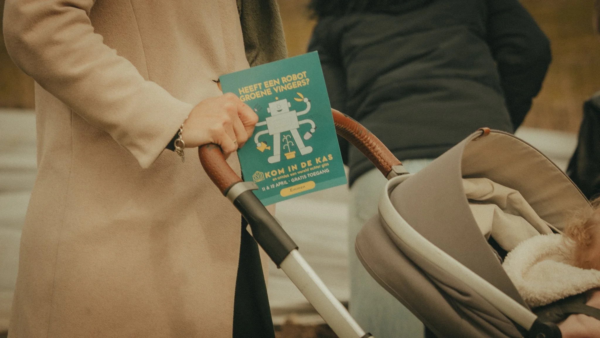 Person holding a pamphlet with a cartoon robot and Dutch text, standing next to a stroller with a dog inside.