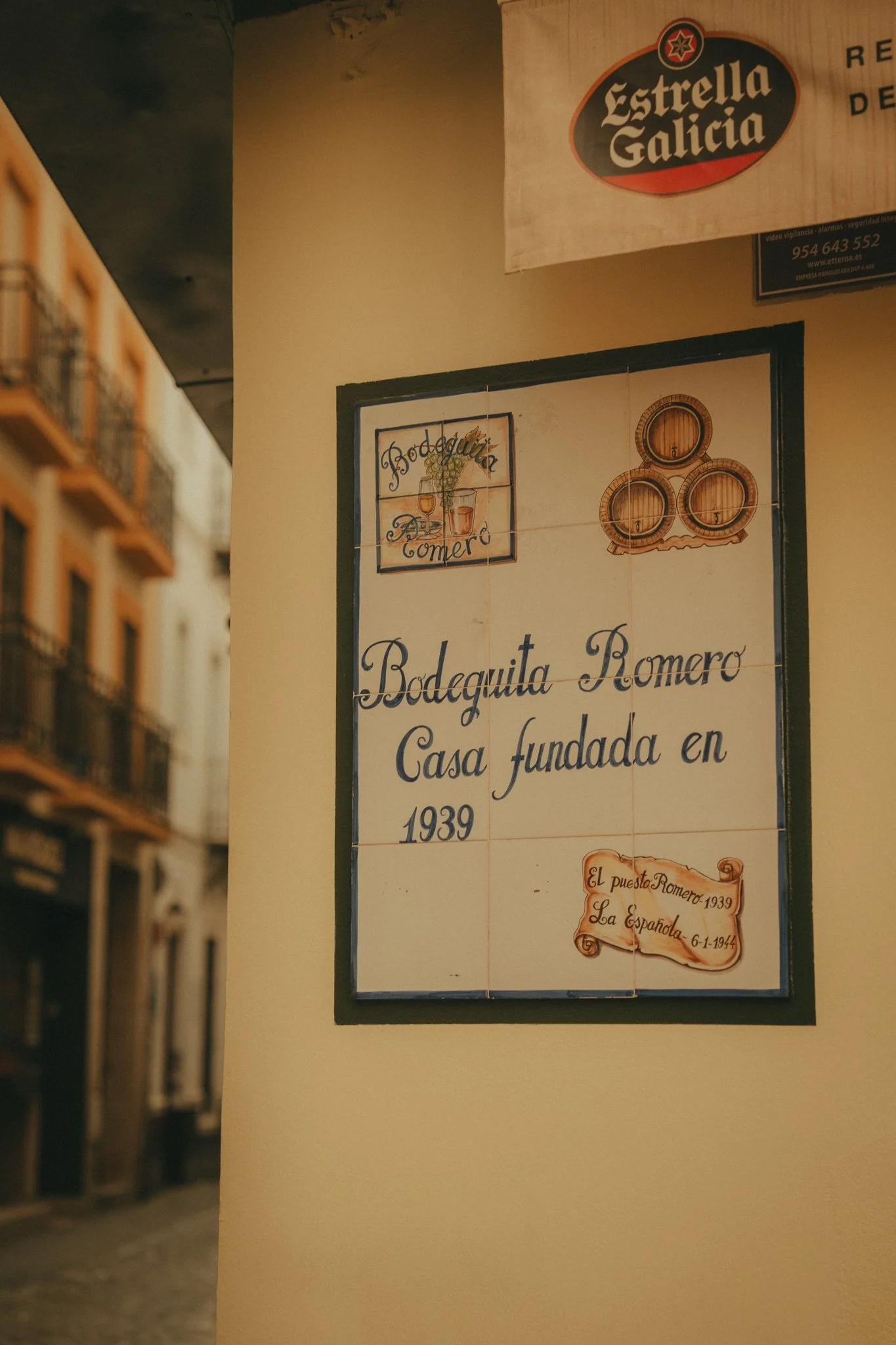 Part of a sign on a wall with text in Spanish, including the name 'Bodeguita Romero,' and illustration of wine barrels and a framed sign with a glass of wine and grapes.