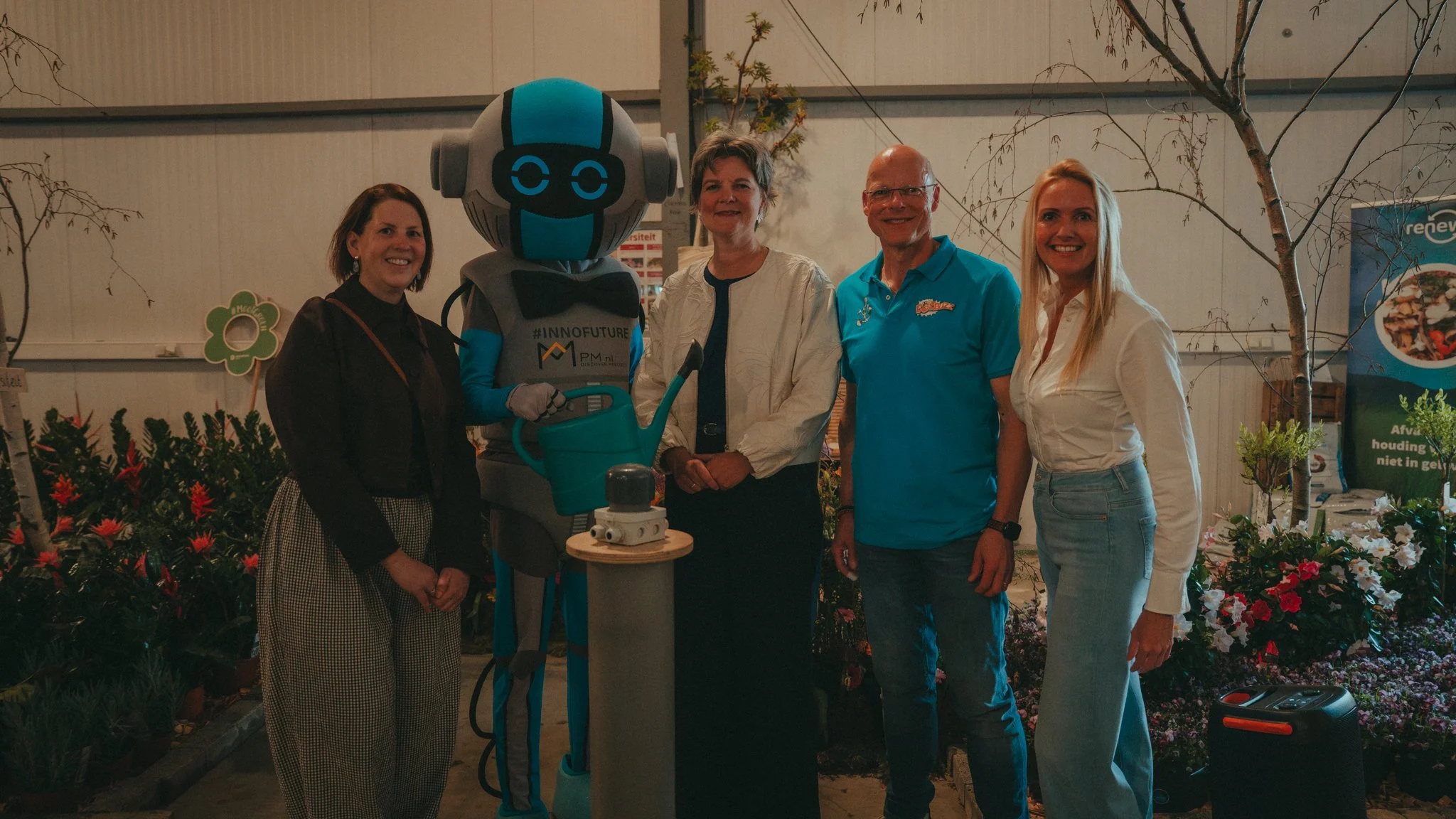 A group of four women and a robot with a digital face, standing together in a garden center with flowers and plants, during evening time.