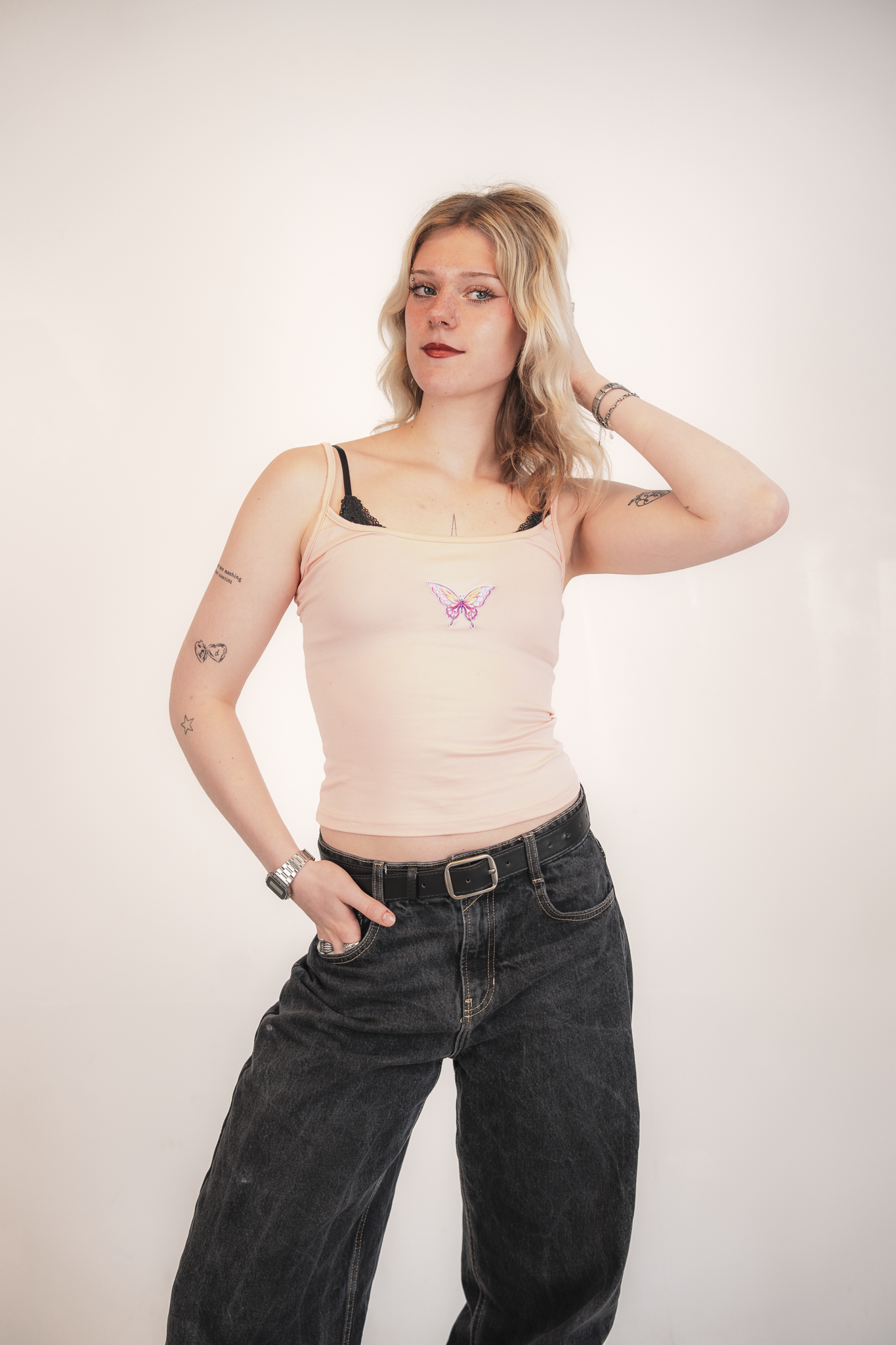 A young woman with blonde, wavy hair and blue eyes poses against a plain white background. She is wearing a light pink tank top with a butterfly design on the front, black bra straps are visible, and she has various tattoos on her arms, including a h