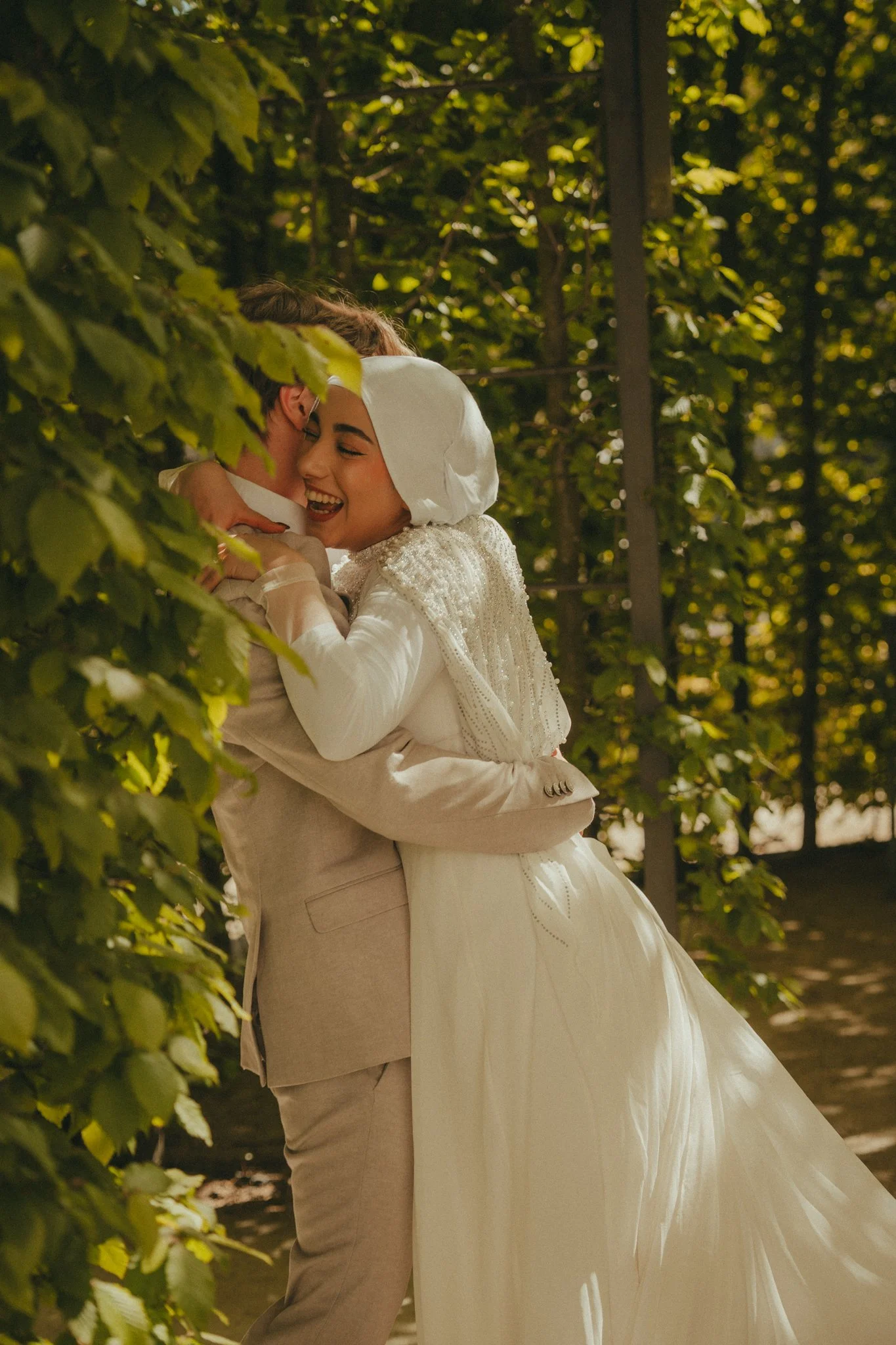 A bride and groom embrace outdoors on their wedding day, surrounded by green trees and sunlight, smiling happily.