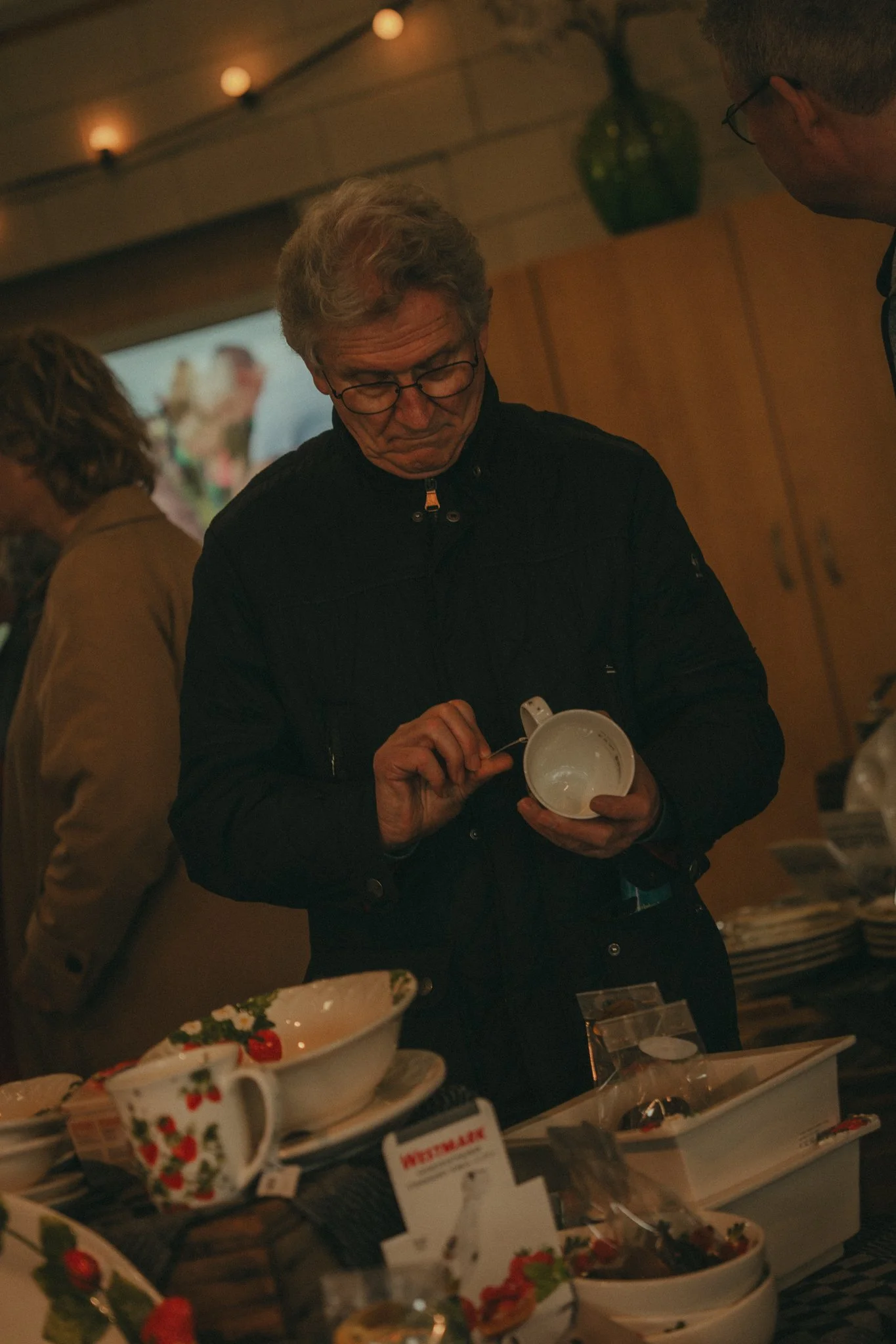 A man in glasses stands at a table filled with bowls of food, holding a white mug and spoon, with a background of a cozy, dimly lit room and festive decor.