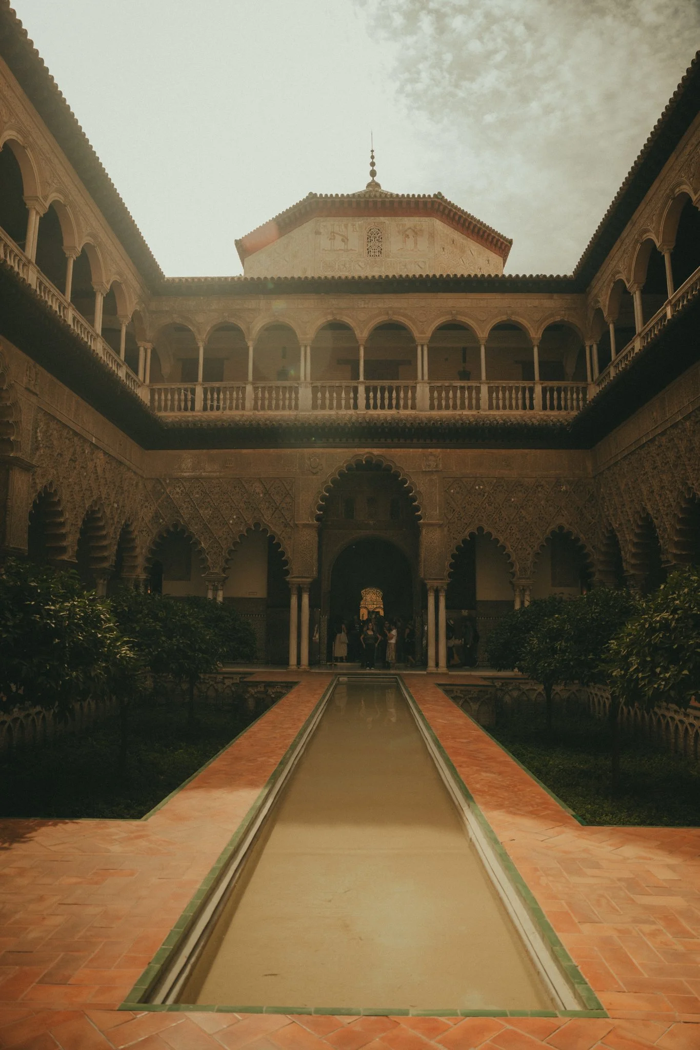 Inner courtyard of a historical building with a central fountain, surrounded by ornate arches and balconies, with a view of the sky and a dome in the background.