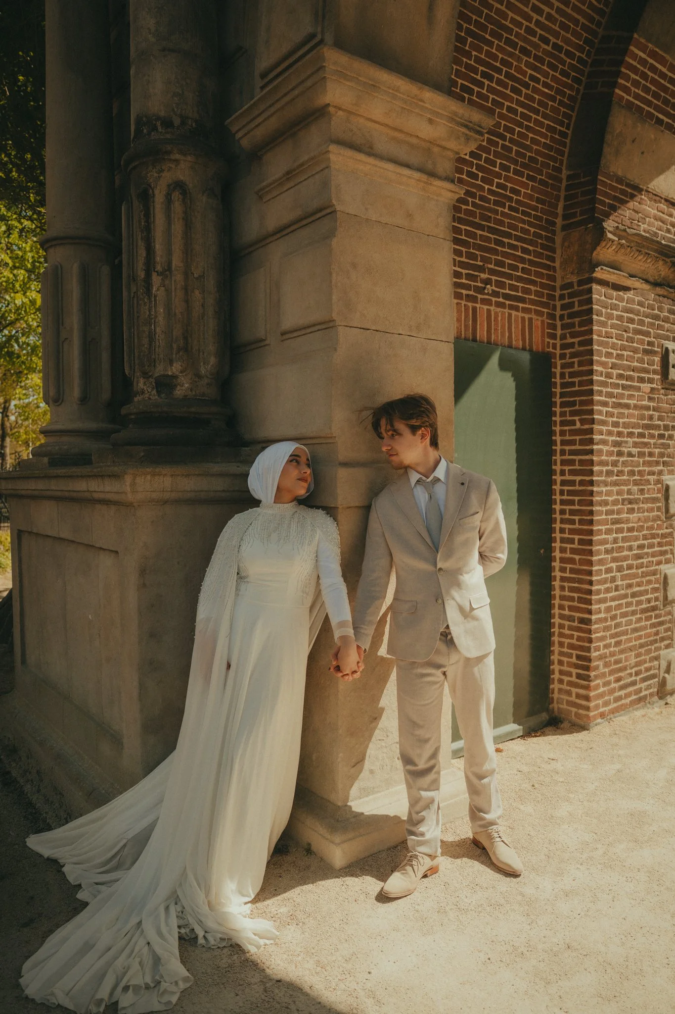 A bride and groom holding hands and looking at each other, standing outside against a historic brick building with large stone columns, during daytime.
