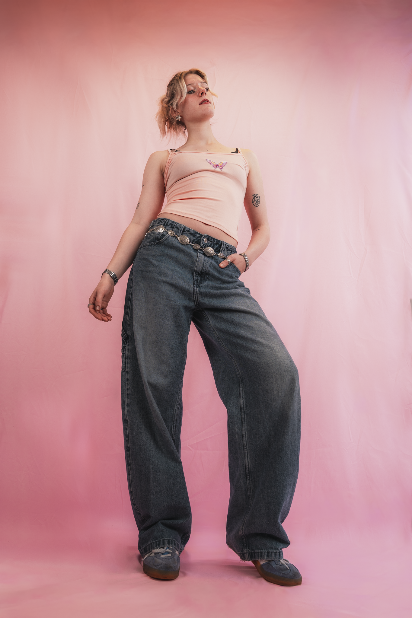 A woman with shoulder-length blonde hair, wearing a pink tank top with a butterfly design, loose dark jeans, and sneakers, standing against a pink background.