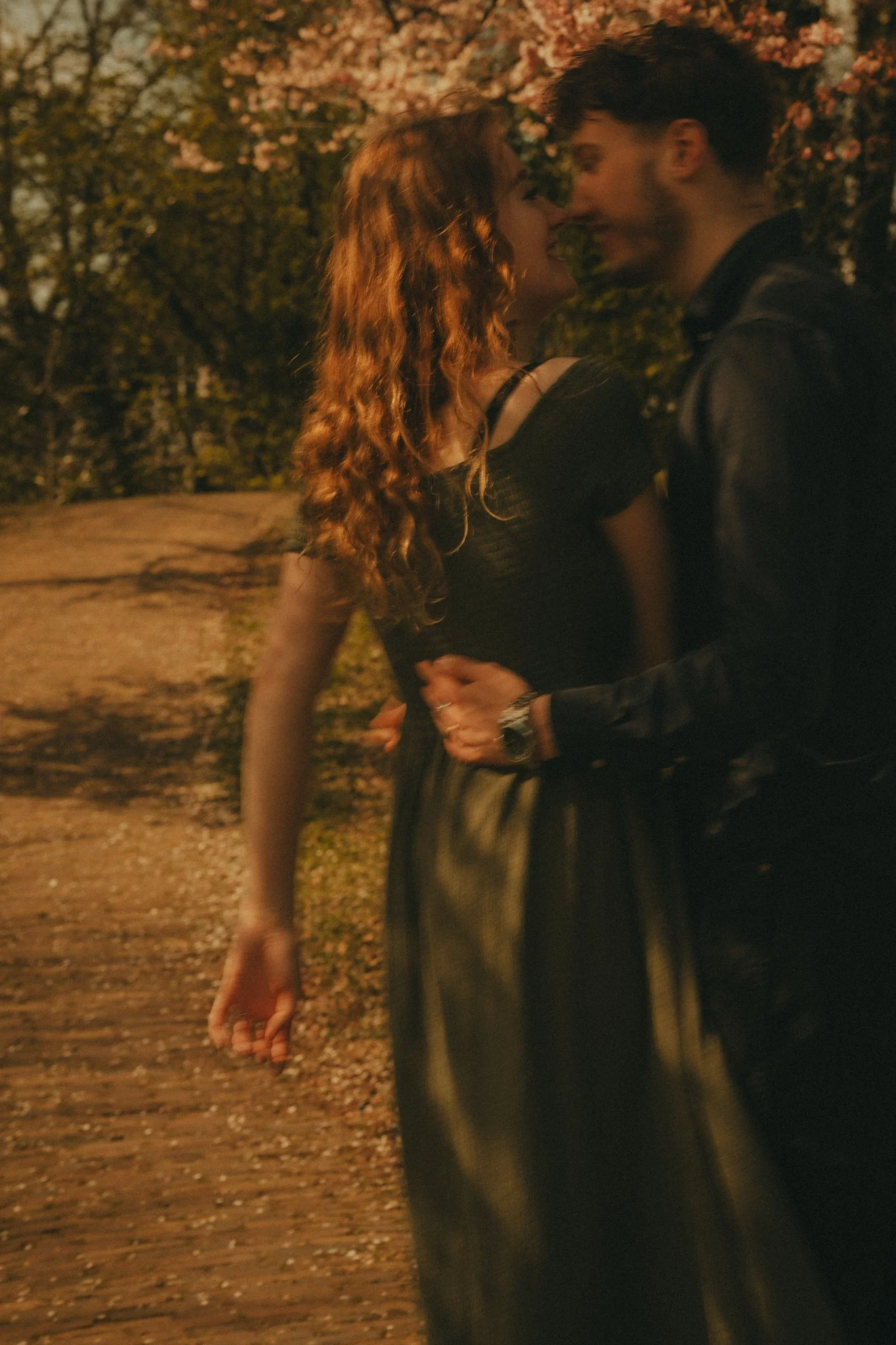 A couple is close together, about to kiss, during sunset in a park with blooming trees and a cobblestone path.