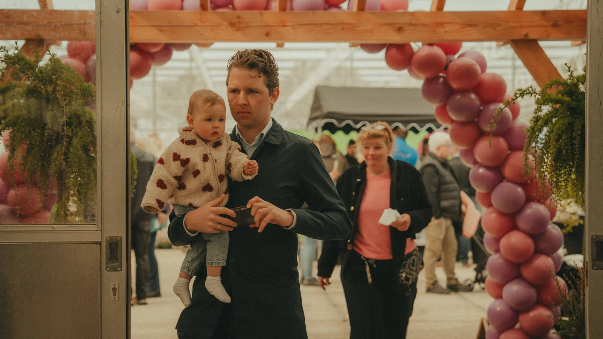 A man carrying a young girl in a cozy jacket through a busy indoor event, with balloons and people in the background.