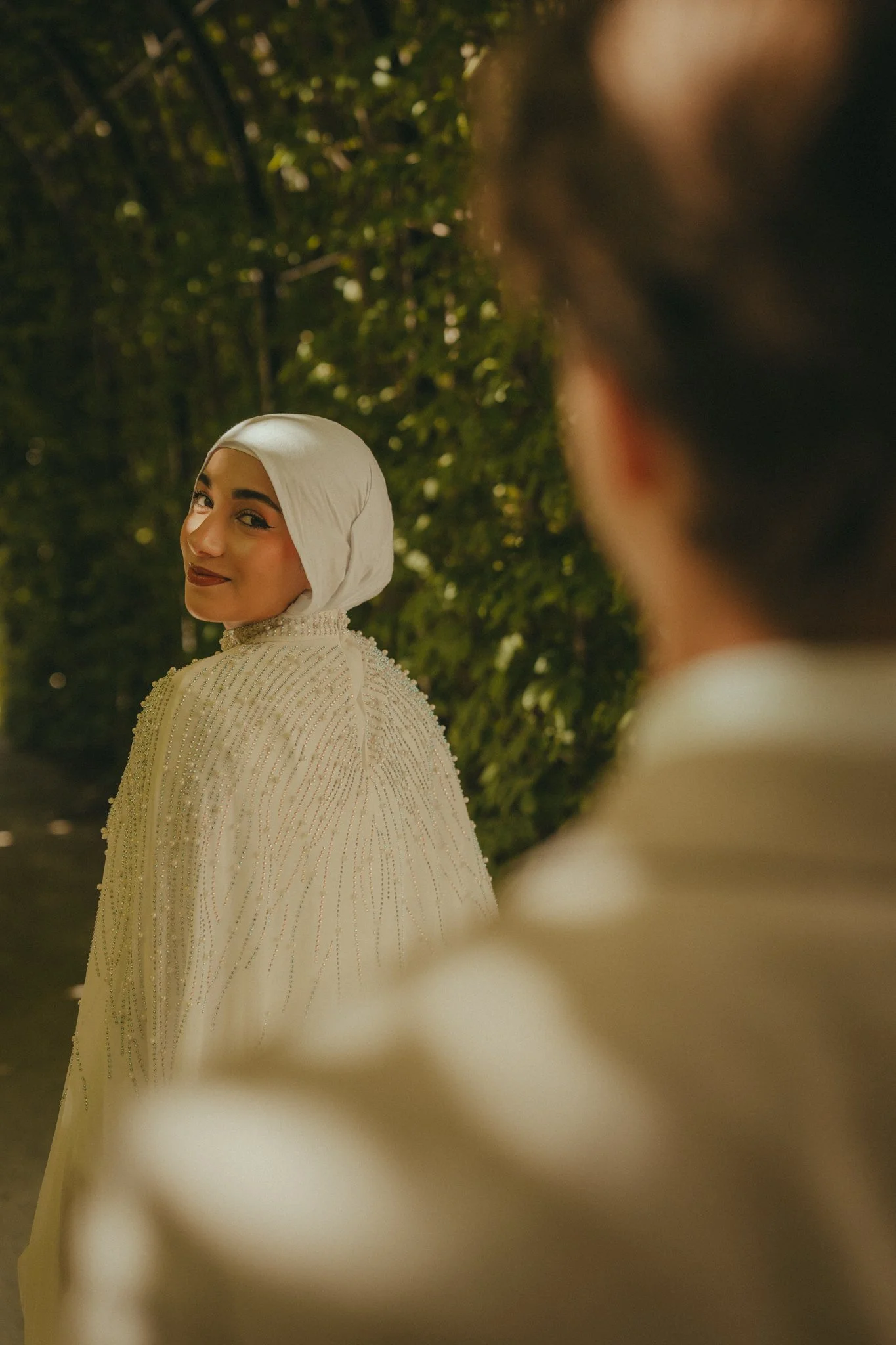 A woman wearing a white hijab and beaded dress smiling at the camera, with a blurred man in the foreground and a green leafy background.