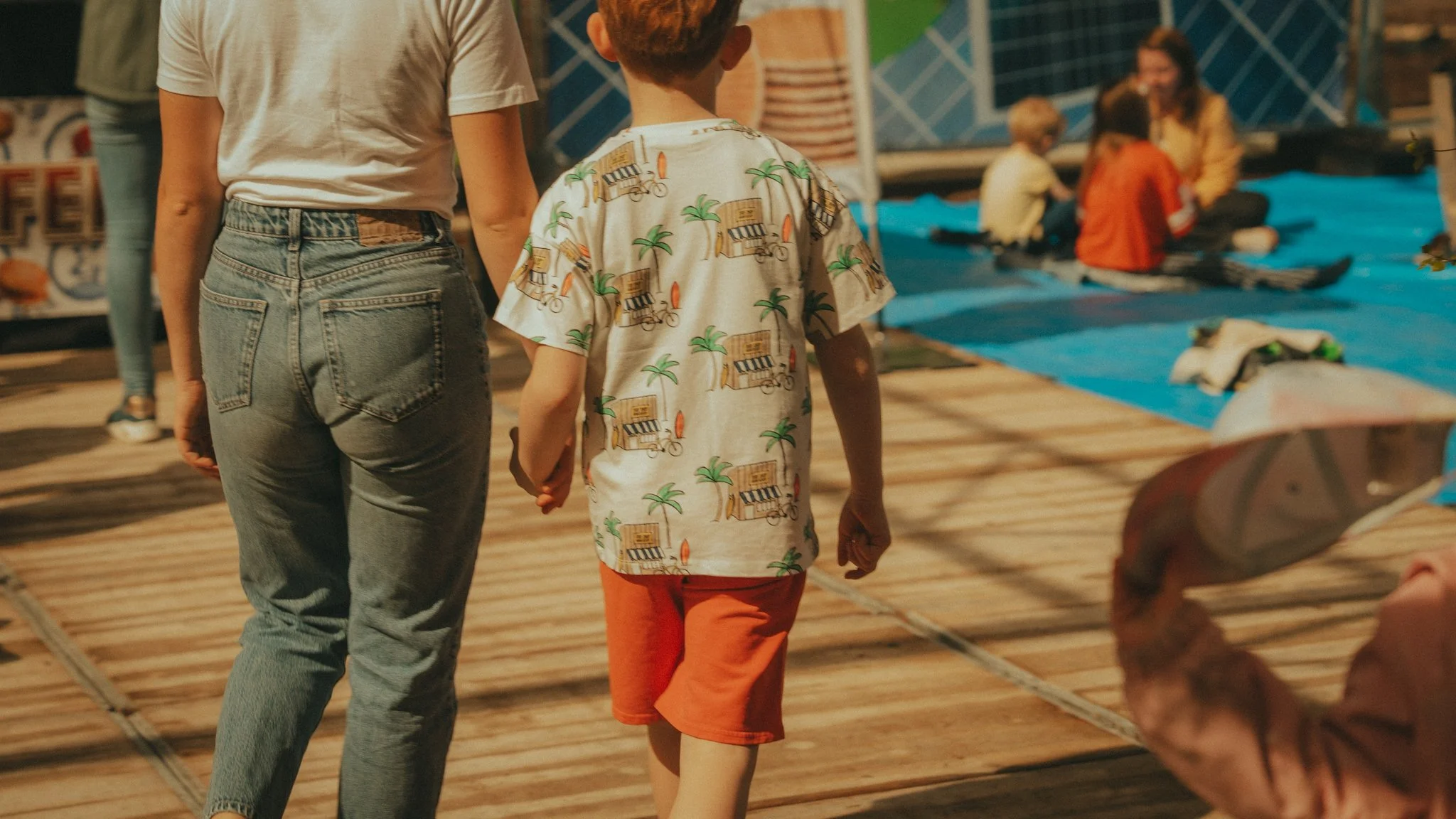 A woman and a young boy holding hands while walking on a wooden pier. The woman is wearing a white t-shirt and jeans, and the boy is dressed in a tropical-themed shirt and orange shorts. In the background, there are children and adults sitting and en