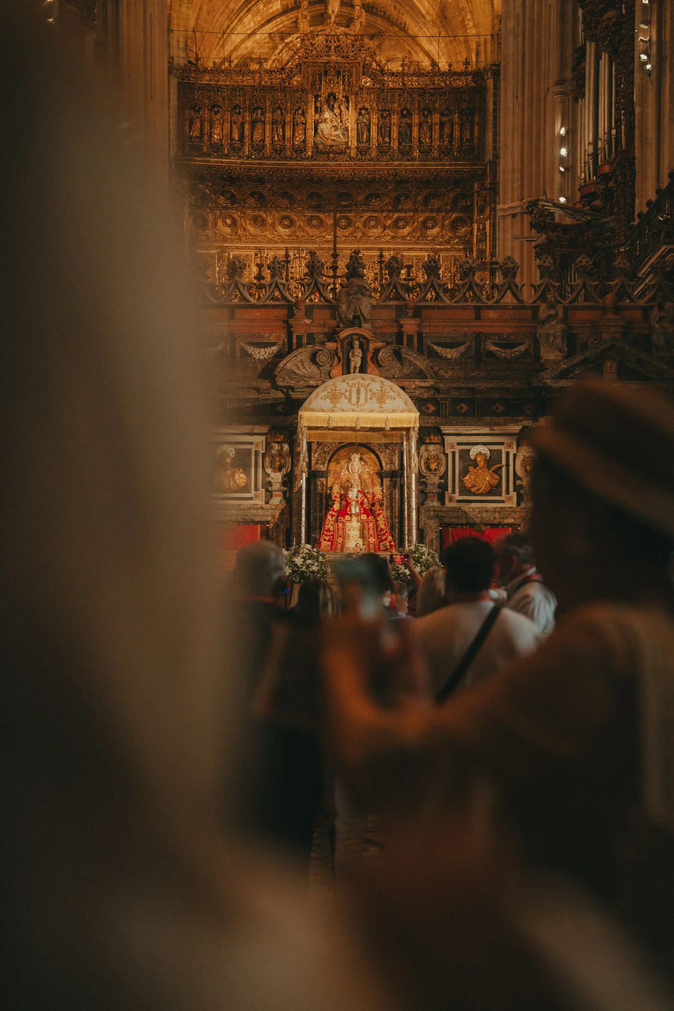 Interior of a grand cathedral with intricate gold and wooden decorations, featuring an altar with a statue of a religious figure, people gathered in worship or attendance.