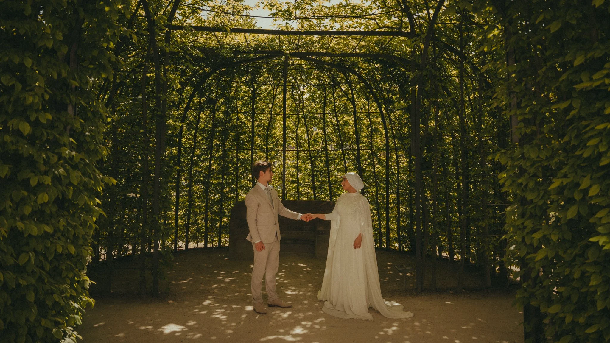 A wedding couple holding hands inside a leafy, archway-like structure with sunlight shining through the trees.