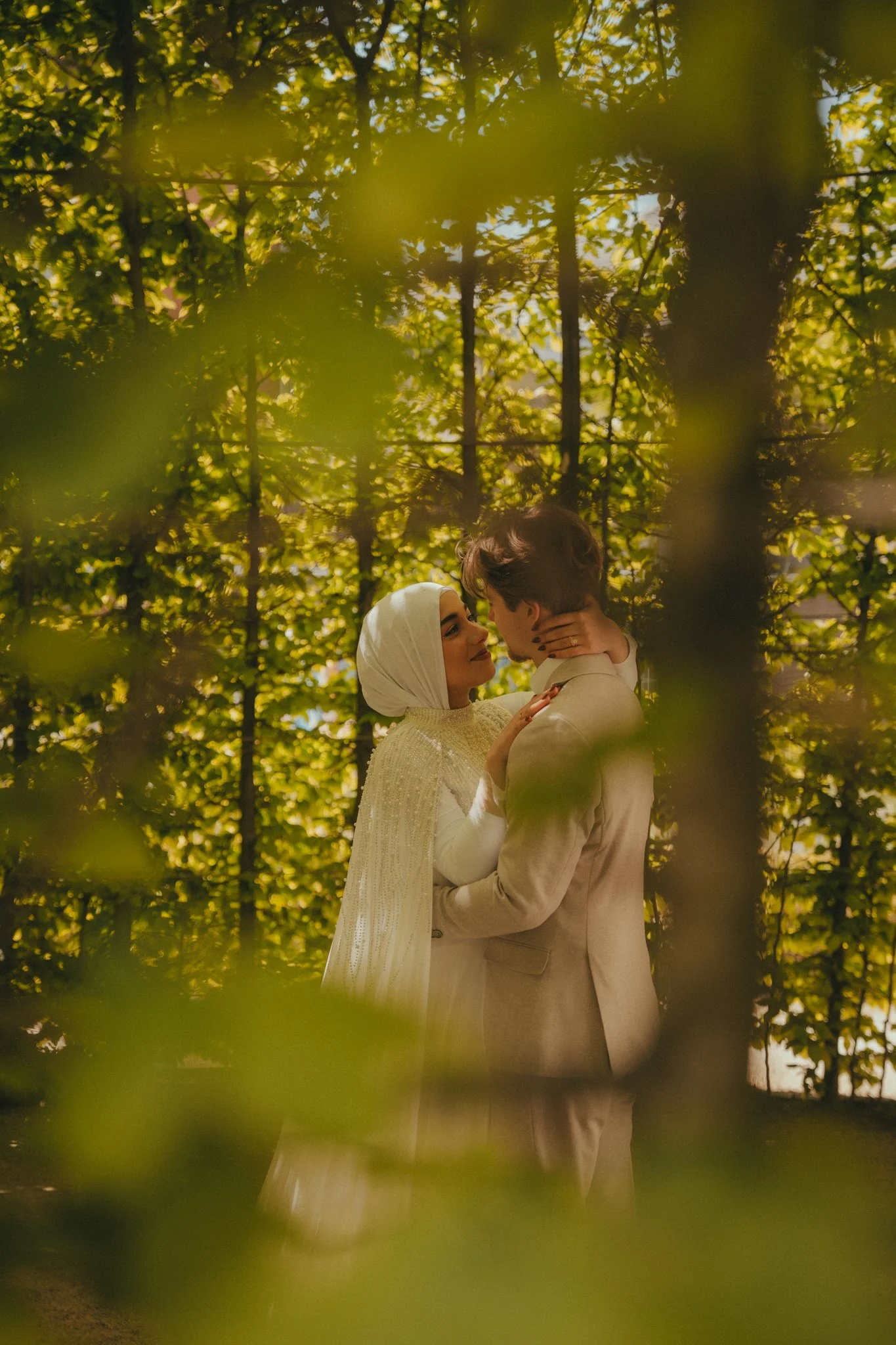 A couple embracing in a lush green outdoor setting, surrounded by leaves and trees, with sunlight filtering through the foliage.