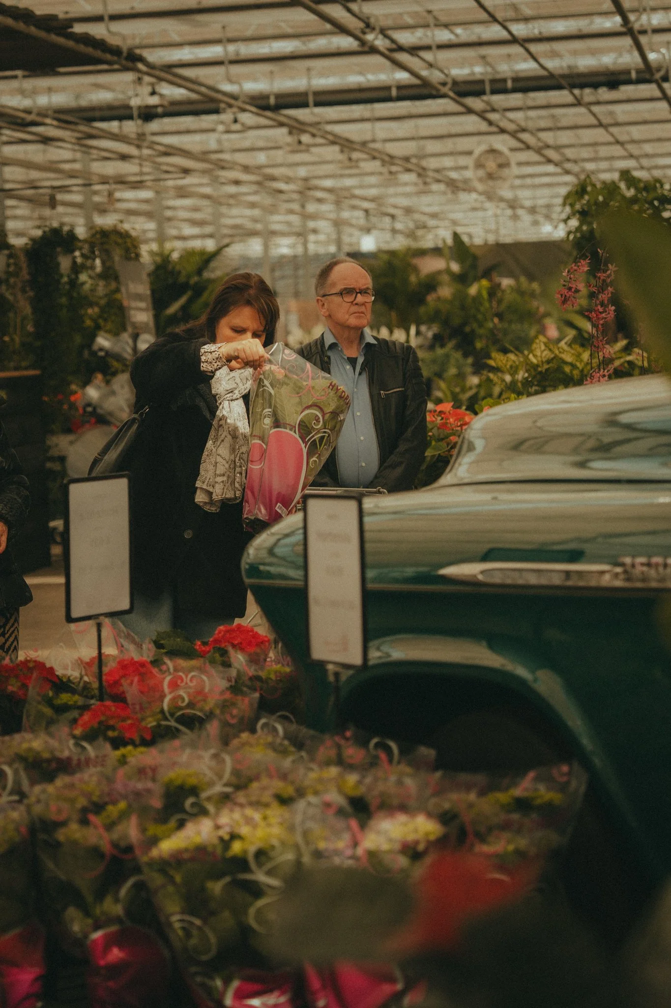 Two people at a flower market, one woman inspecting a bouquet and a man standing with arms crossed, surrounded by flowers and plants under a glass roof.