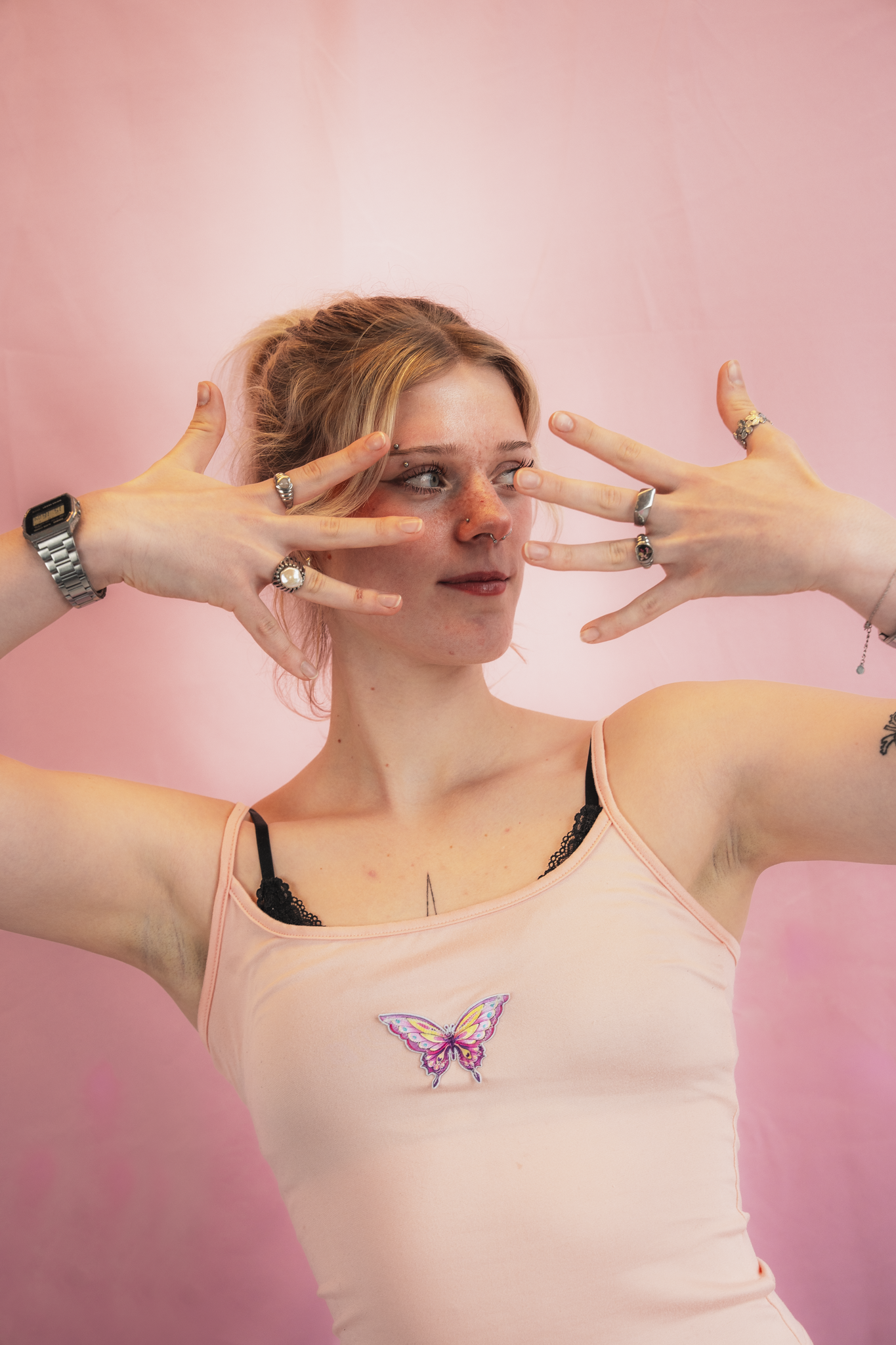 Young woman with light brown hair poses against a pink background, wearing a light pink spaghetti strap top with butterfly embroidery and multiple rings and jewelry on her fingers.
