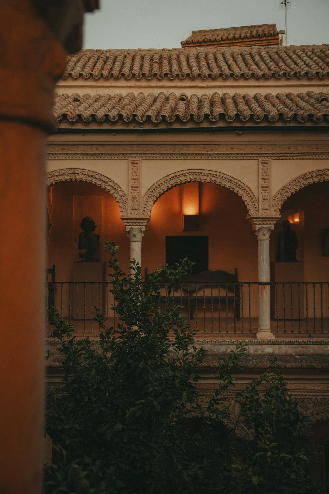 View of a building with a tiled roof, ornate columns, and arches, with statues inside the building, and greenery in the foreground.