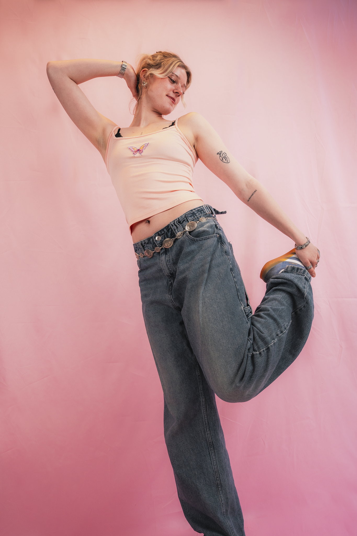 A young woman with tattoos wearing a pink tank top and jeans, striking a playful pose with one leg bent and one arm behind her head, standing against a pink background.