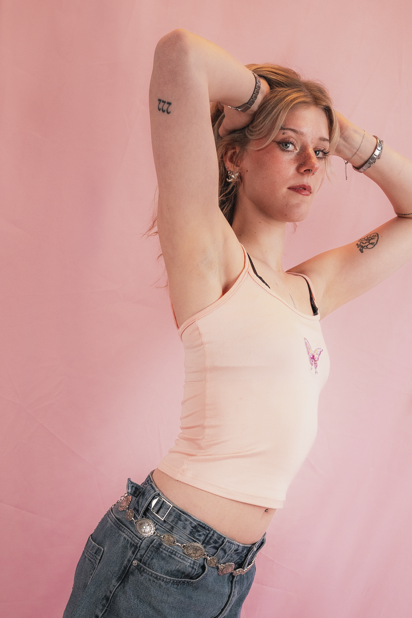 A young woman with blonde hair, blue eyes, and freckles, standing against a pink background. She has tattoos on her arms, wears a necklace, earrings, bracelets, and a chain belt with coins. She is wearing a peach-colored tank top with a butterfly emb