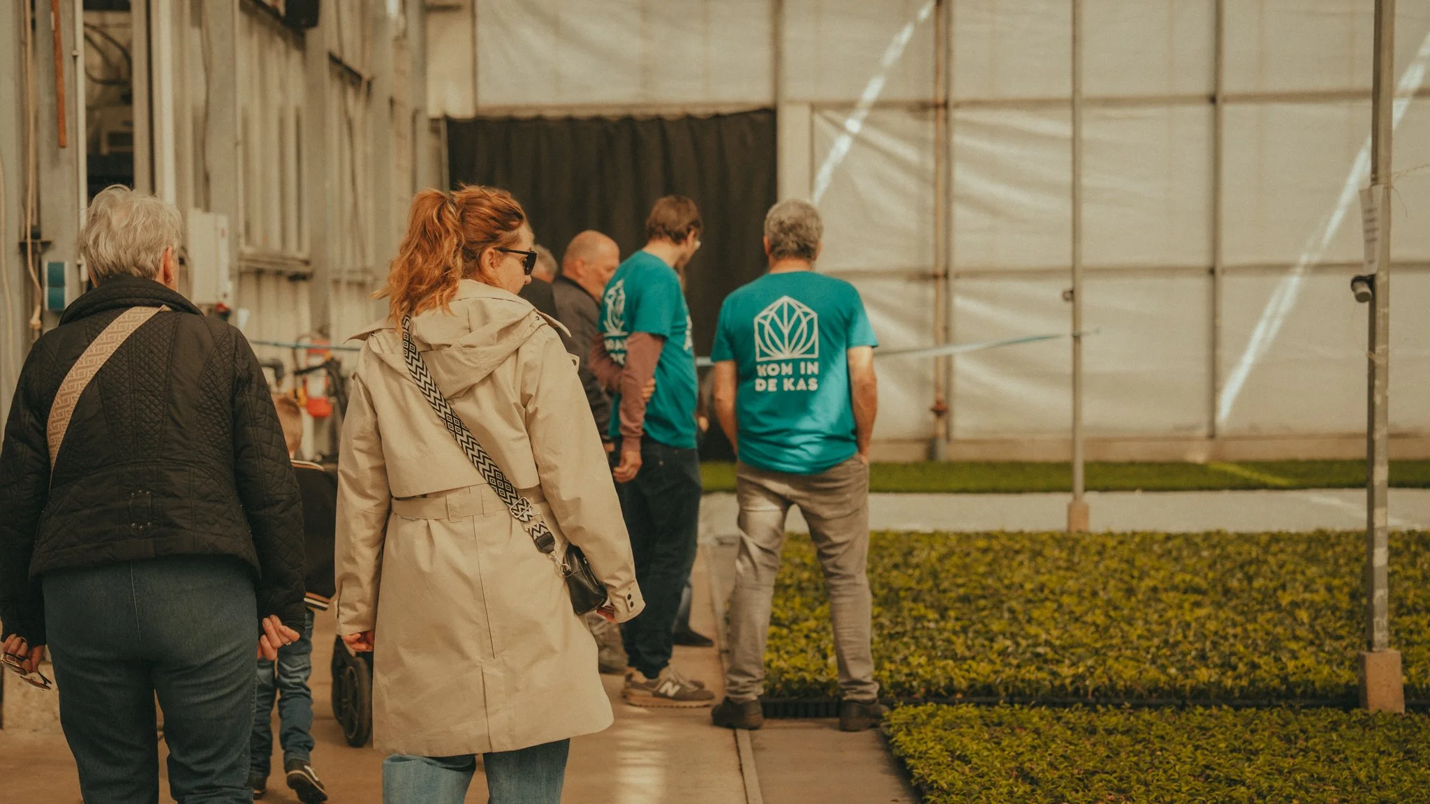 A group of people standing in line inside a building, possibly waiting for an event or entrance, with some individuals wearing teal shirts and casual clothing. There is a large structure with metal framing and curtains in the background.