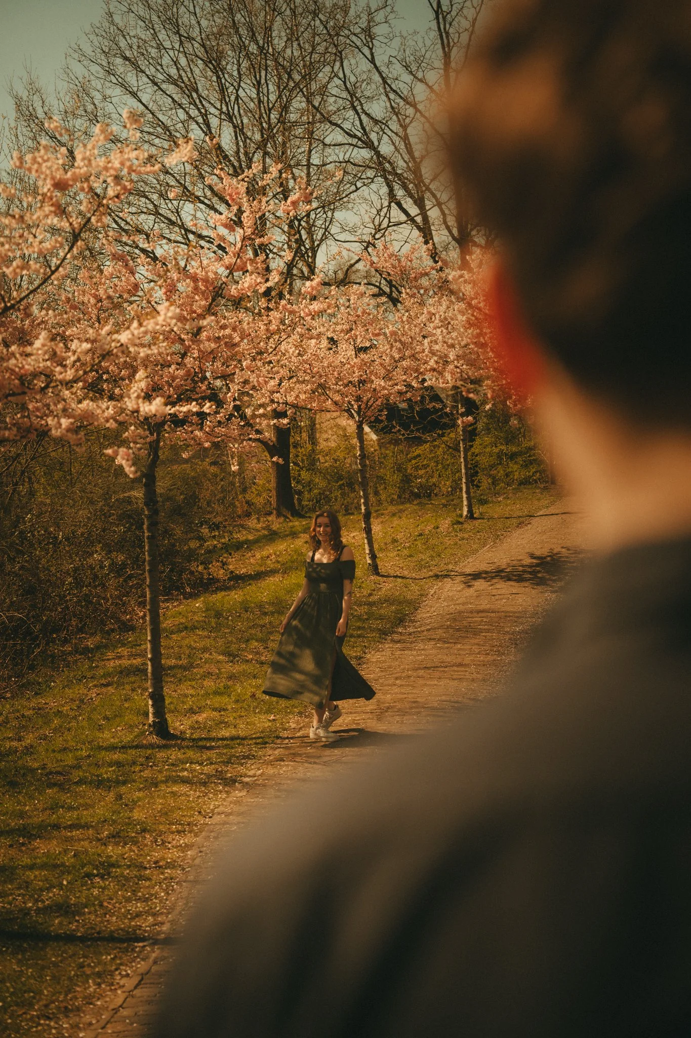 A young woman in a black and green dress walking along a park path surrounded by blooming cherry blossom trees, with a blurry object partially blocking the right side of the photo.
