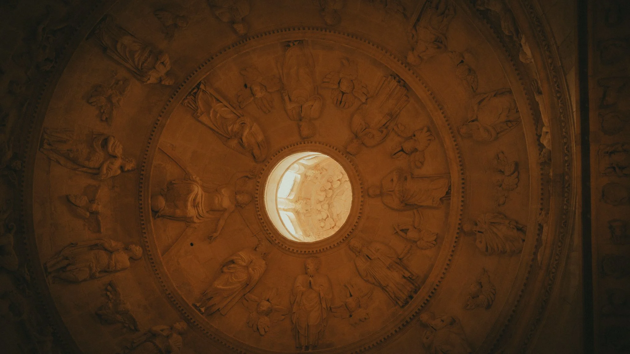 Ceiling view of a domed architectural structure with intricate sculptures and a central opening allowing light in.