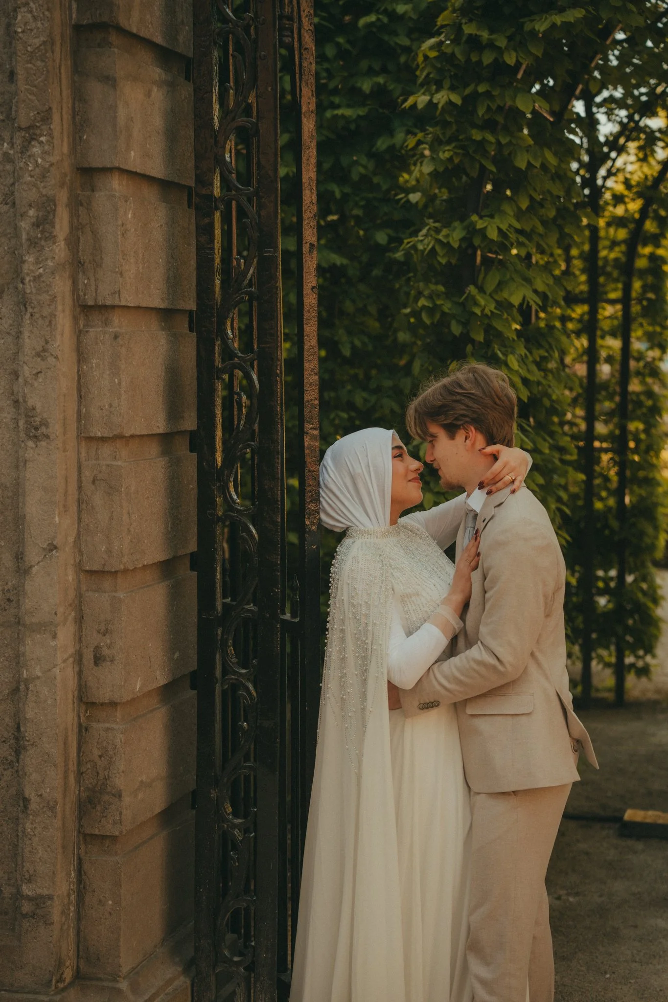 A couple stands close together outdoors, embracing and gazing into each other's eyes amidst greenery and sunlight, with a stone wall and metal fence in the background.