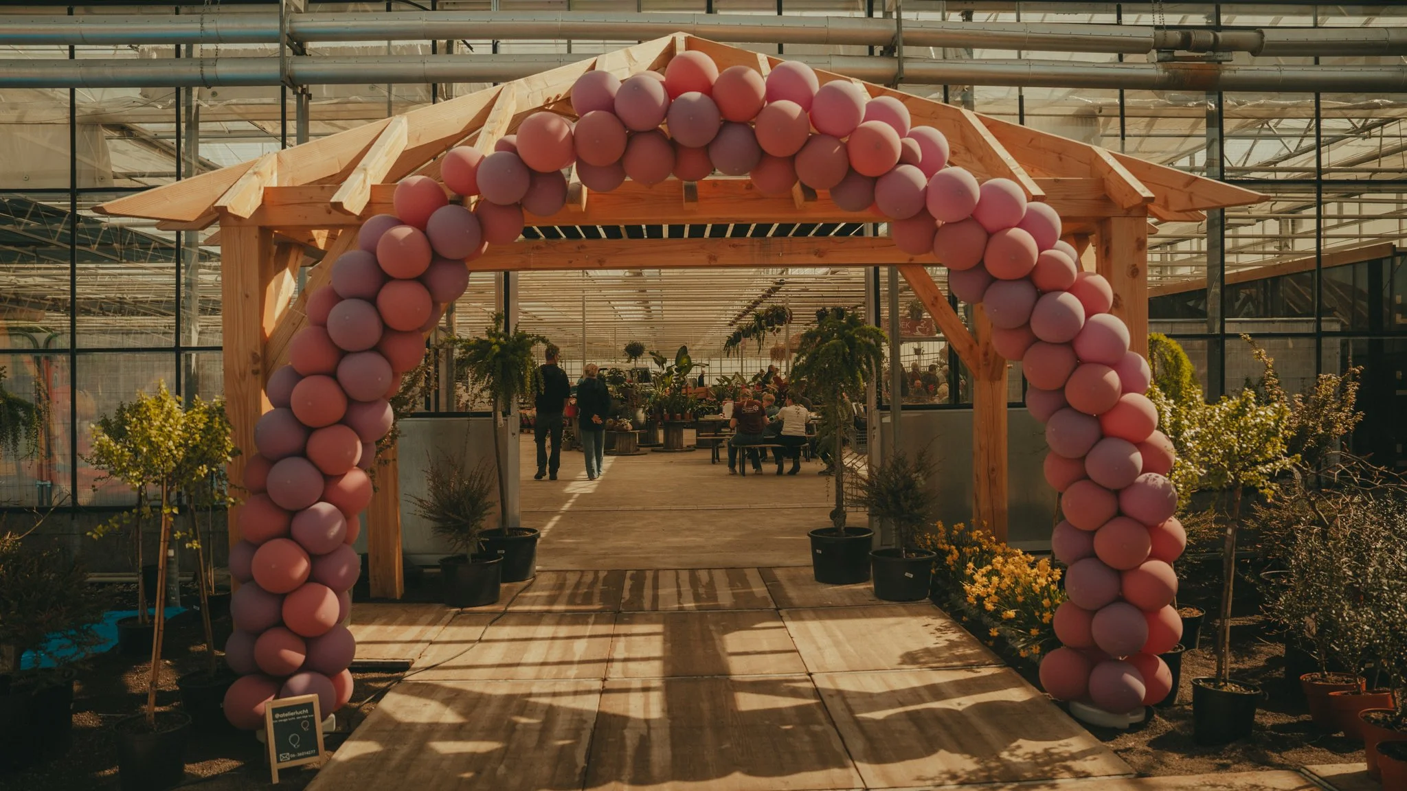 Wooden archway decorated with grass and pink balloons at the entrance of a greenhouse or flower shop, with a few people inside.