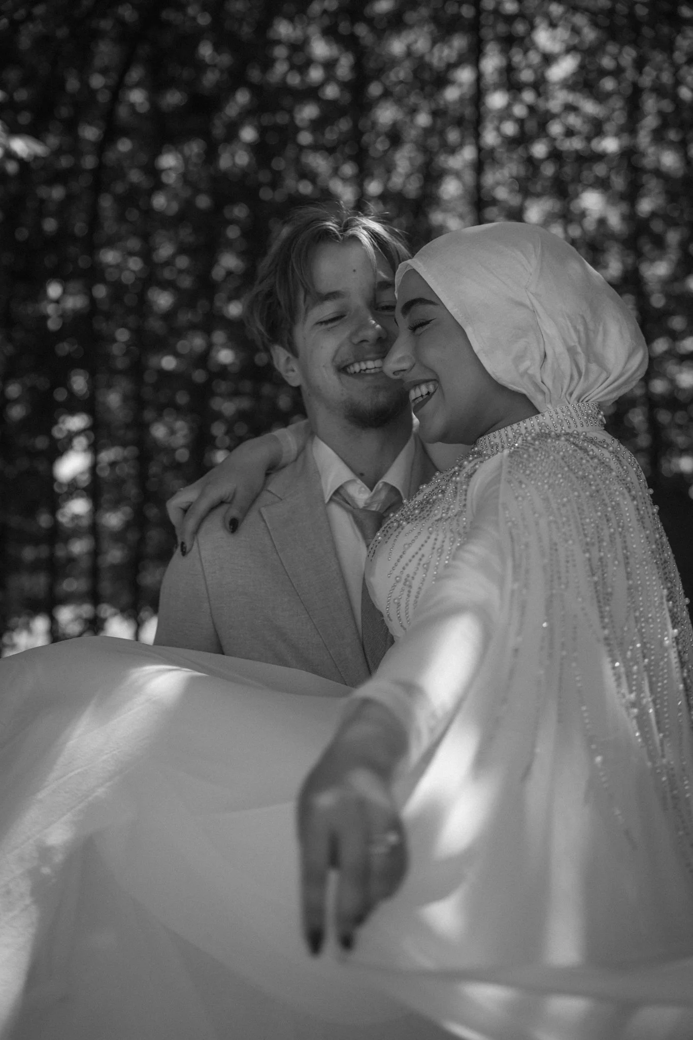 Black and white photo of a smiling young man and woman hugging outdoors, with blurred trees in the background, as the woman takes a selfie.