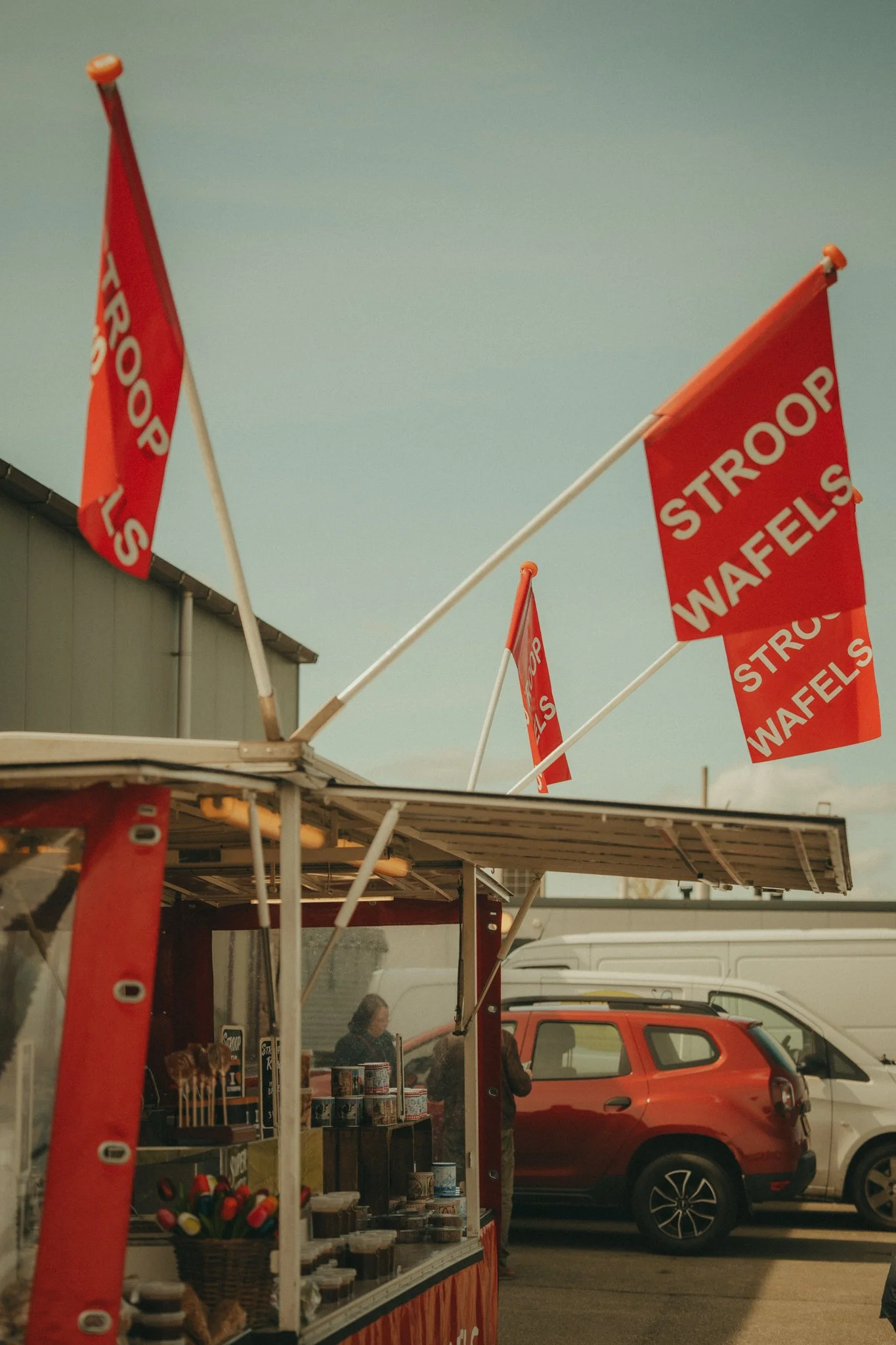 Red flags with white text reading "Strap Waffles" flying above a stall at an outdoor event, with a food stand, grilled items, and parked cars visible in the background.