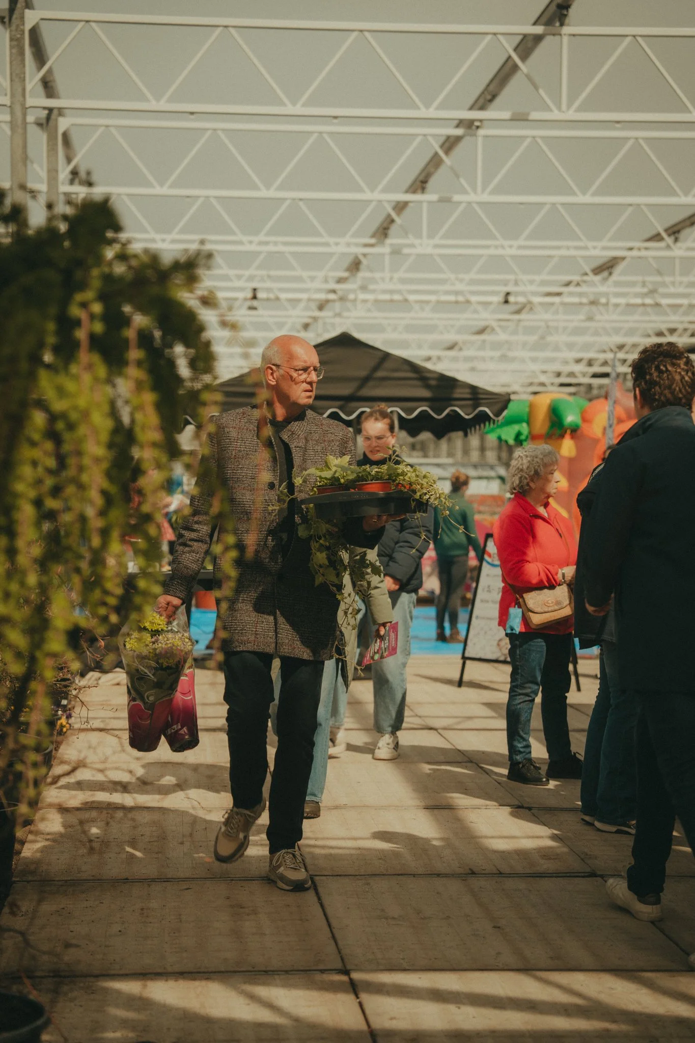 People shopping at an outdoor market under a white metal framework with a black canopy and colorful balloons in the background.