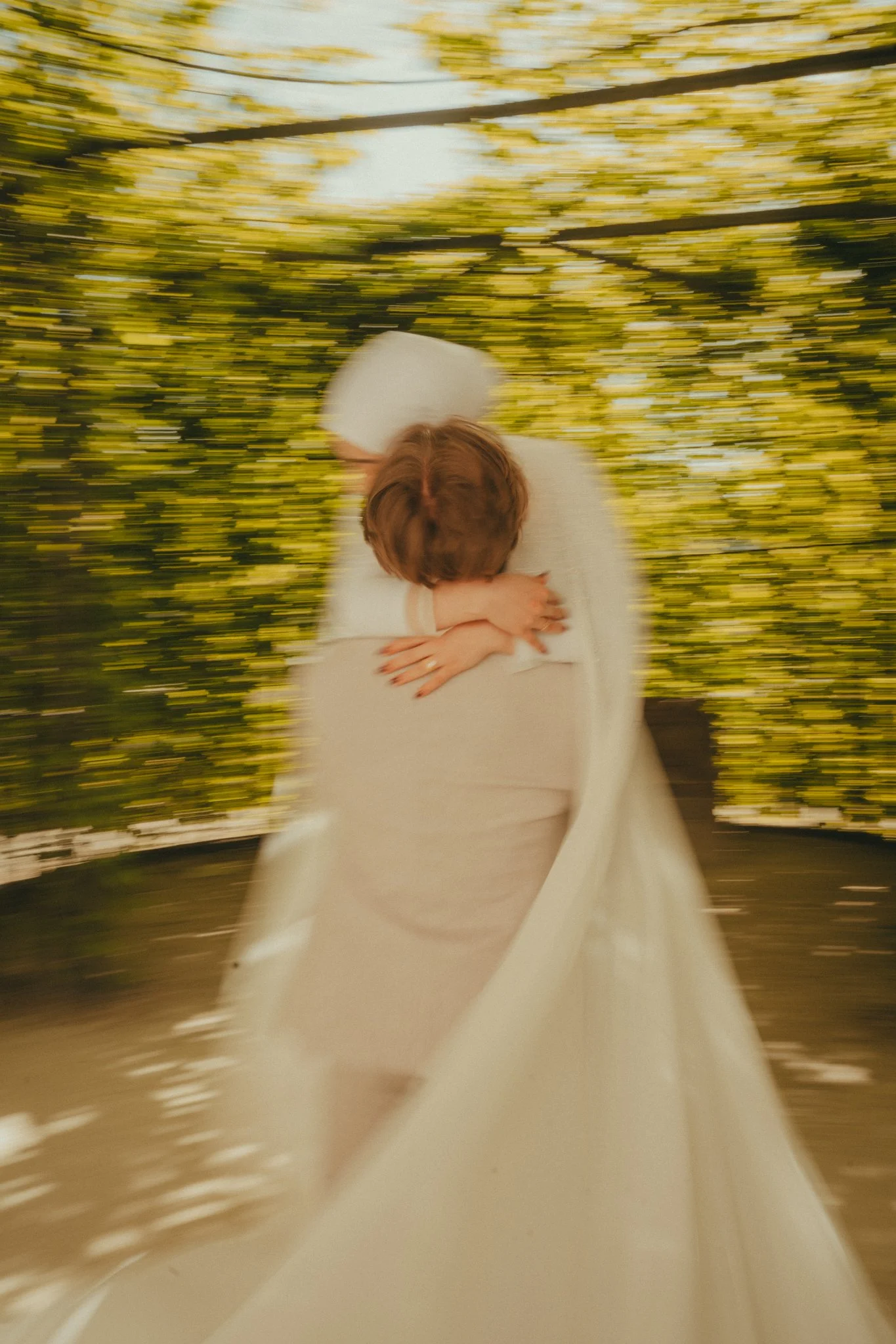 A person in a white wedding dress hugging another person wearing a white head covering, with a background of yellow and green trees, creating a motion blur effect.