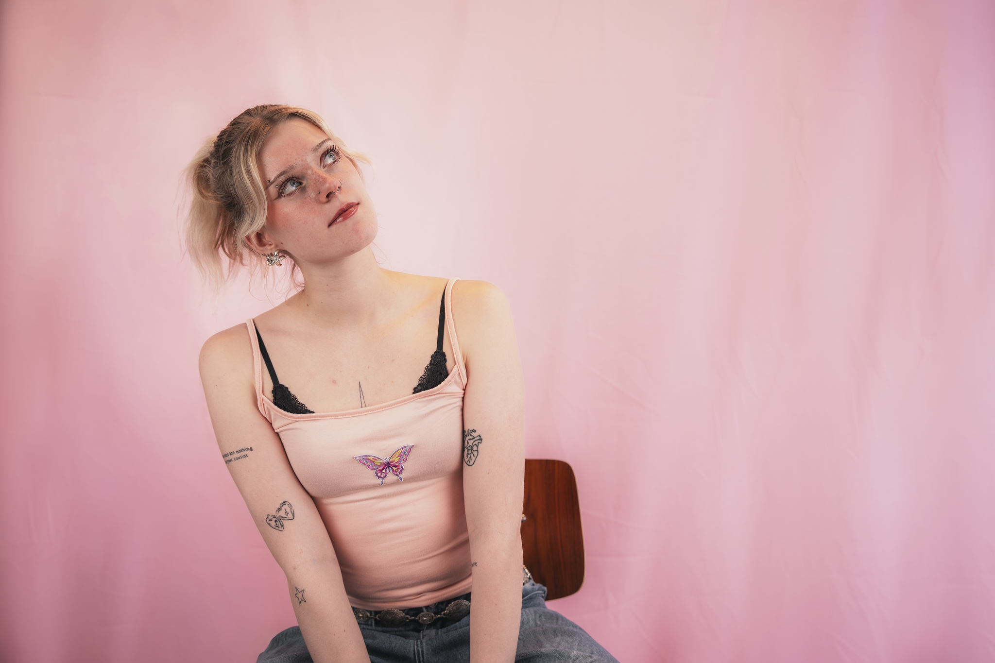 A young woman with short blonde hair, freckles, and multiple facial piercings, sitting against a pink background. She is wearing a pink tank top with butterfly and tattoo designs, and looking upward with a thoughtful expression.