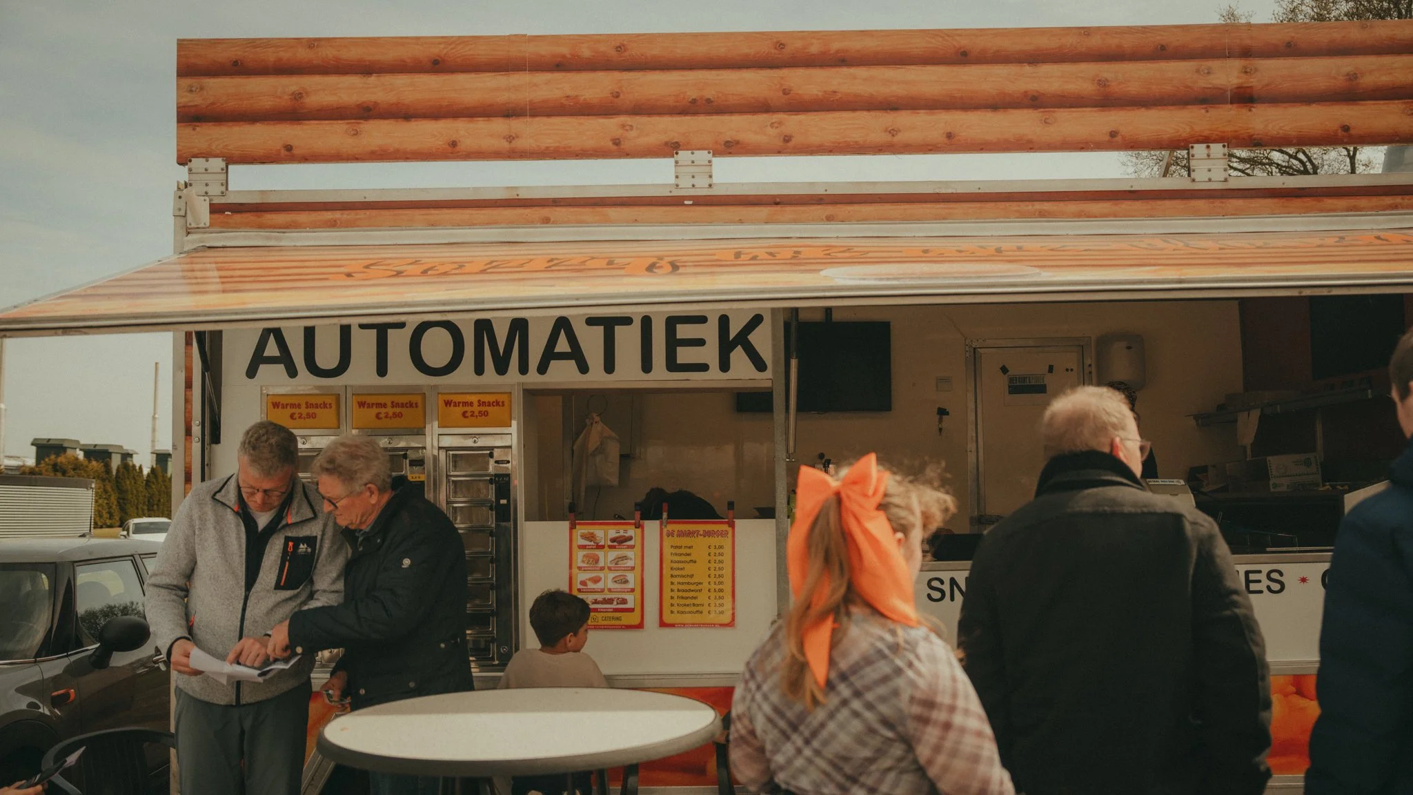 People standing at a food stand with a sign that reads 'AUTOMATIEK', selling hot snacks in a parking lot, with a menu displaying prices.