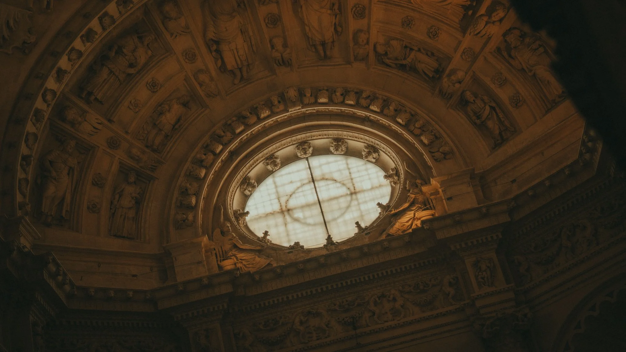 Interior view of a grand architectural dome with detailed sculptures and a large circular skylight at the top, illuminated by natural light.