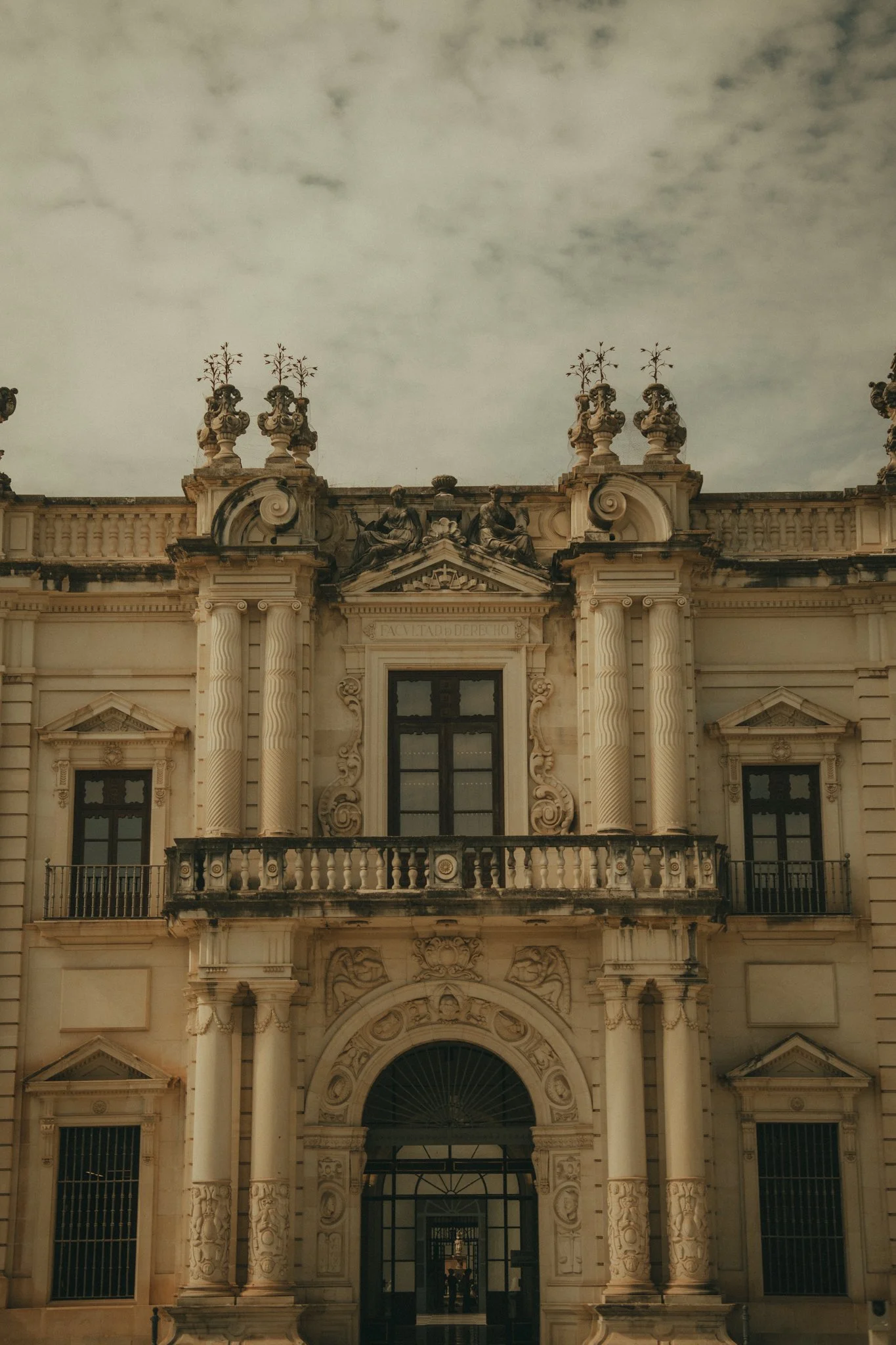 Facade of an ornate historic building with detailed stone carvings, columns, and statues on the roofline, under a cloudy sky.