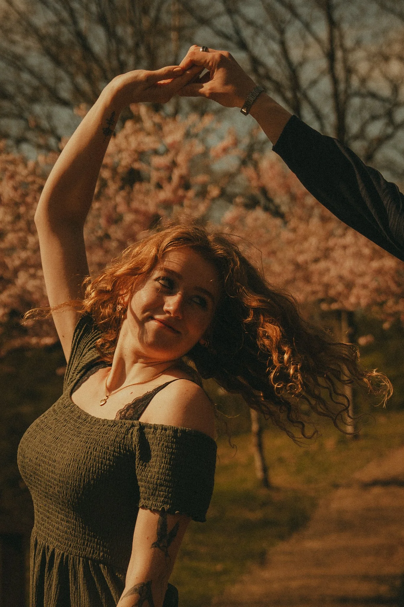 A woman with curly red hair dancing outdoors at night, with another person's hand touching hers, in a park with trees and pink blossoms in the background.