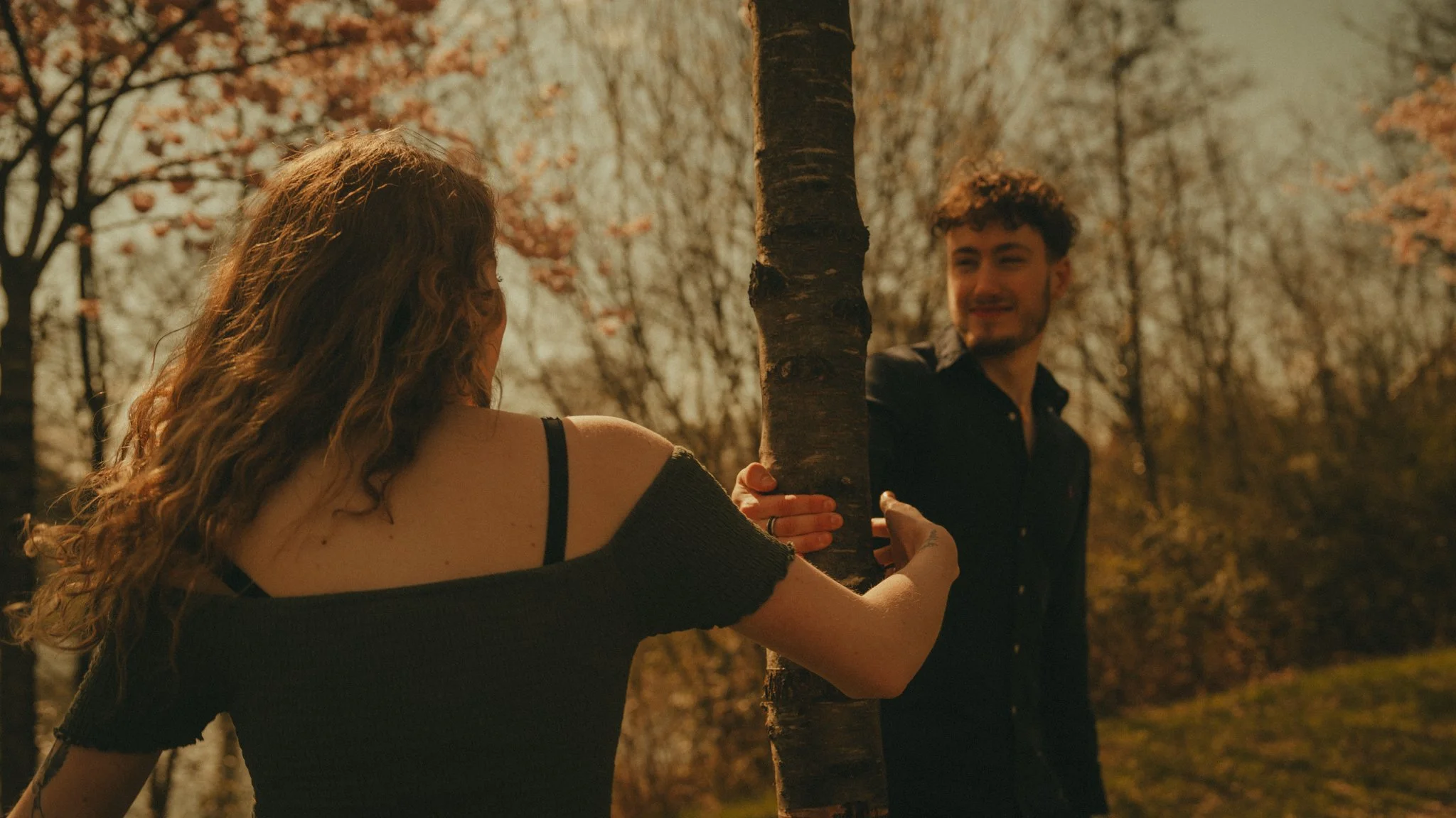 A woman with long curly hair and a man with short hair are outdoors in autumn, near a tree, sharing a moment and holding hands.
