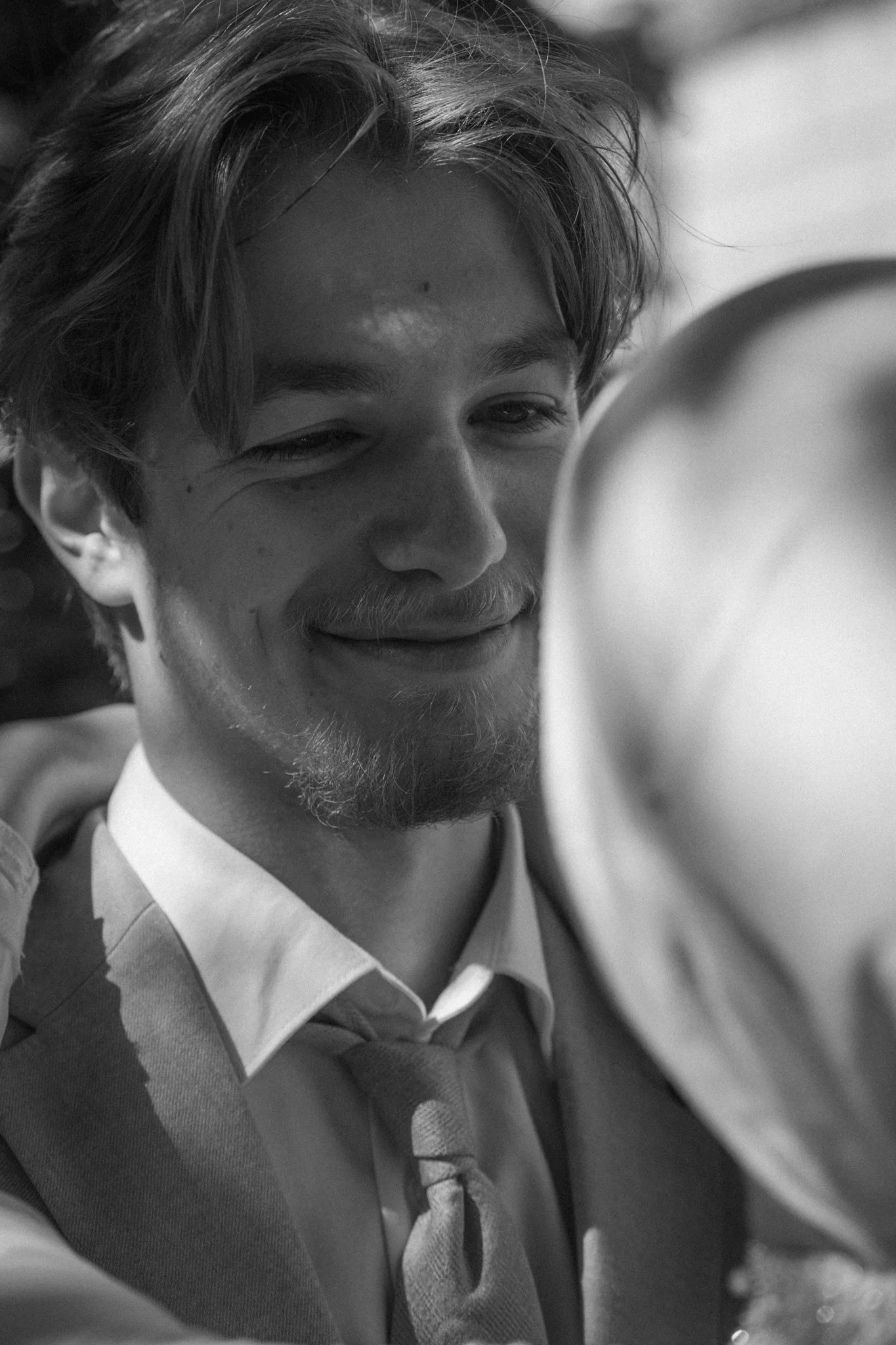Close-up black and white photo of a smiling young man with wavy hair, wearing a collared shirt and a sweater with a tie visible, and blurred figures in the background.