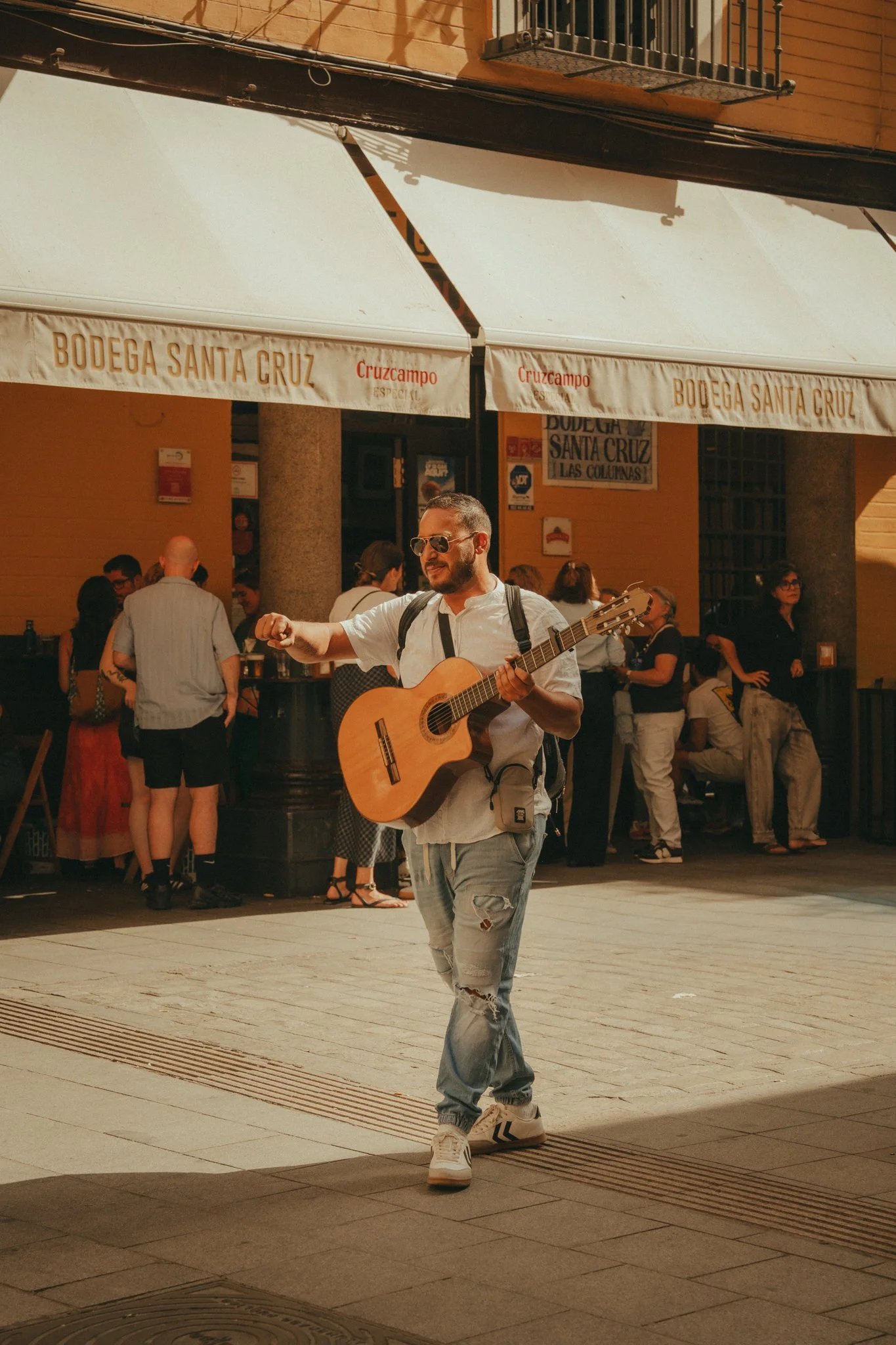 A man wearing sunglasses and a white shirt playing guitar while walking on a busy street outside a bar or restaurant with a crowd of people in the background.